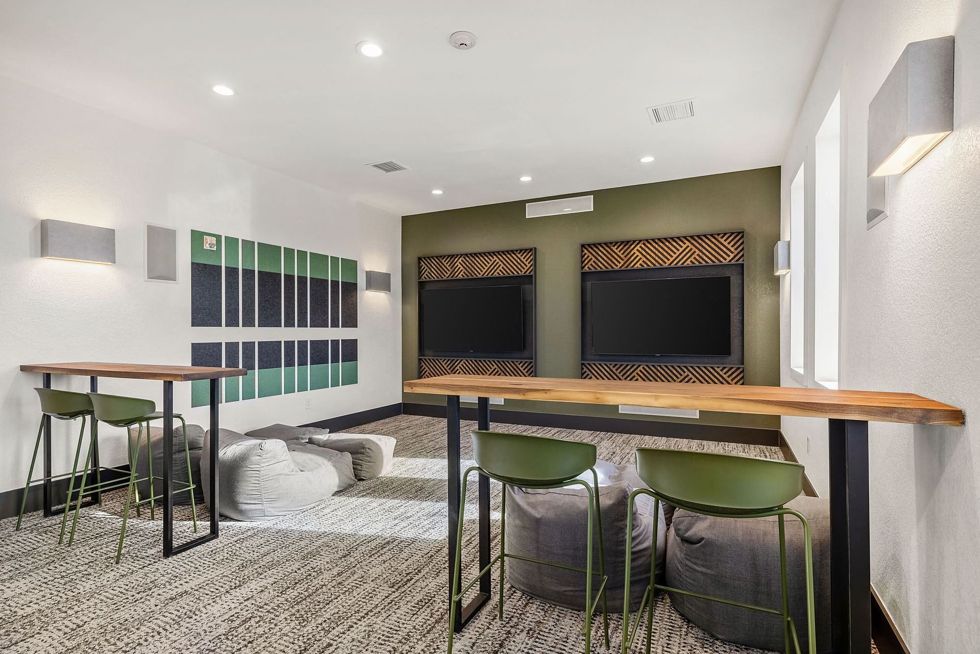 Indoor community lounge with two wall TVs, a long wooden counter, green stools, and bean bag seating.