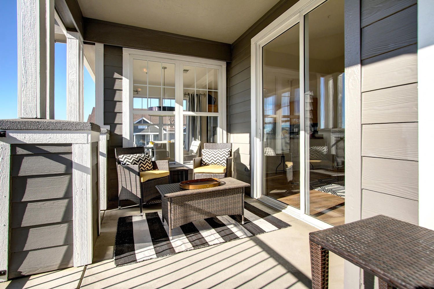 Outdoor apartment balcony with wicker chairs, a coffee table, and a striped rug near sliding glass doors.