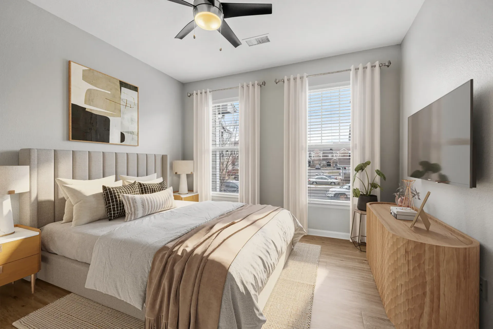 Bright bedroom with beige upholstered bed, two nightstands, dresser with TV, and large windows with white curtains.