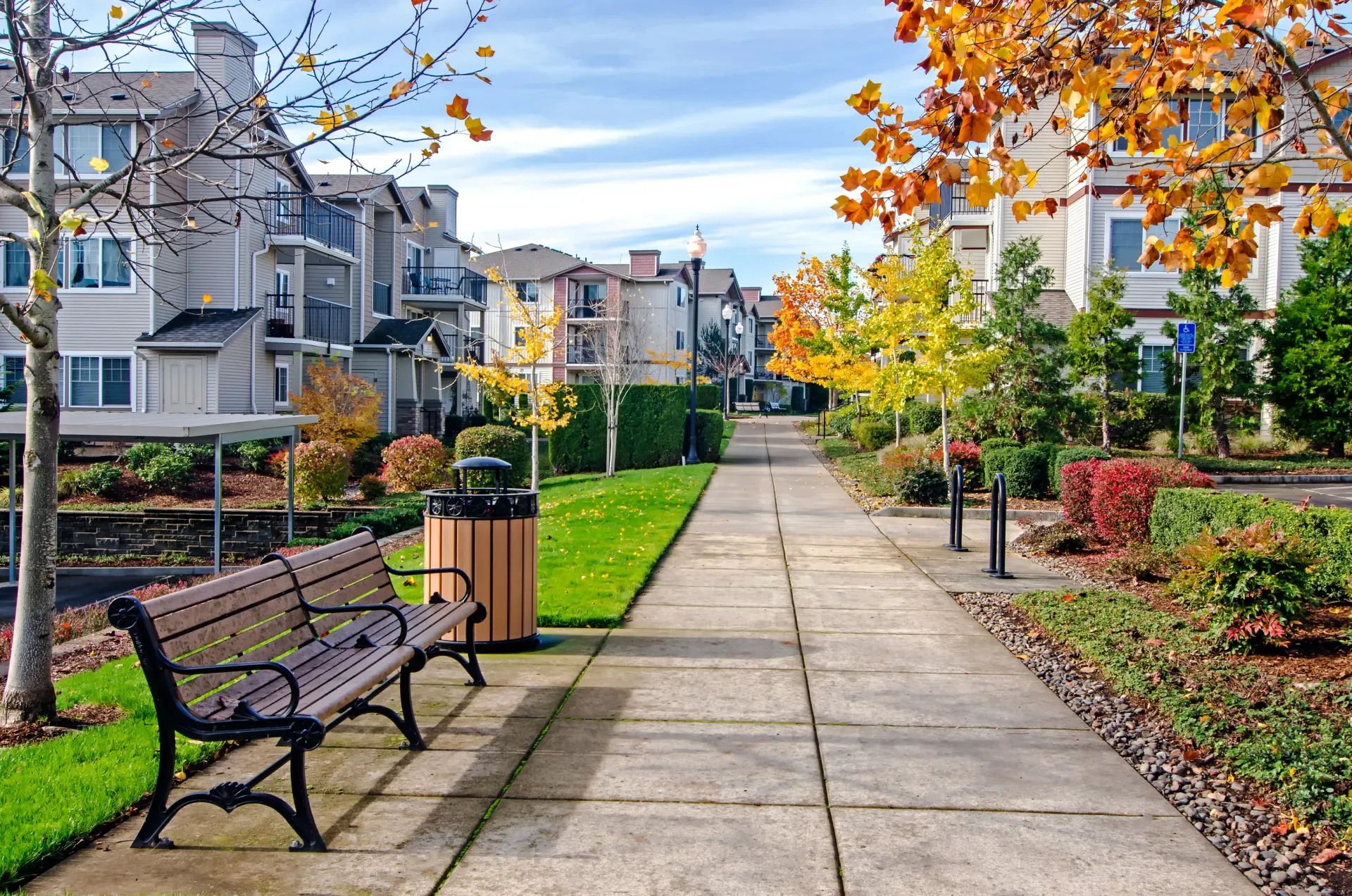 Wide concrete path through an apartment community with benches, lampposts, and fall-colored trees.
