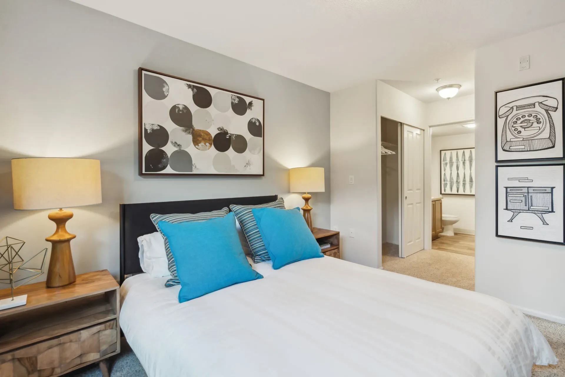 Bedroom in an apartment with a neatly made bed, two wooden nightstands, blue pillows, and wall art.