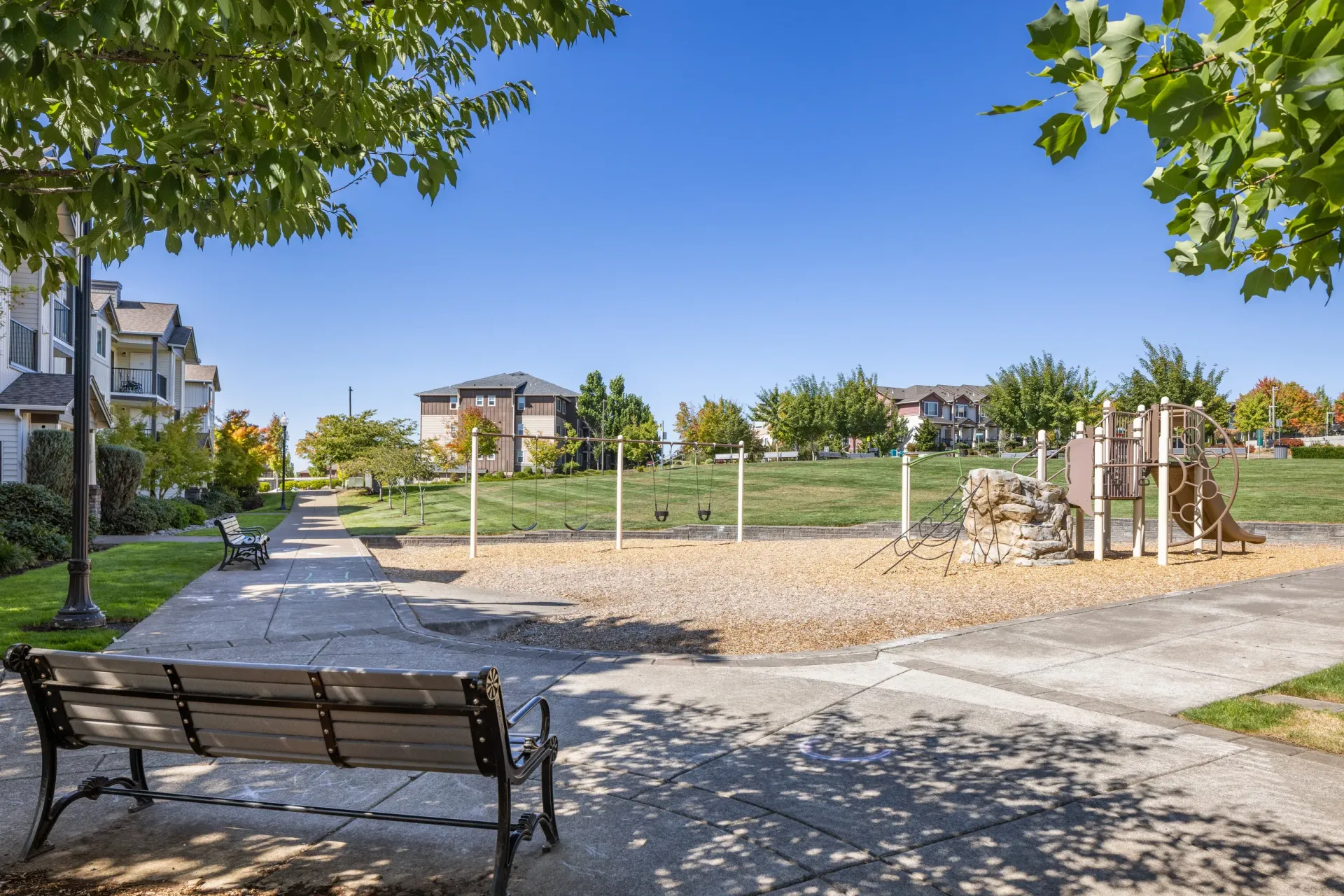 Playground with swings and a slide in a residential apartment community, with benches and paved paths.