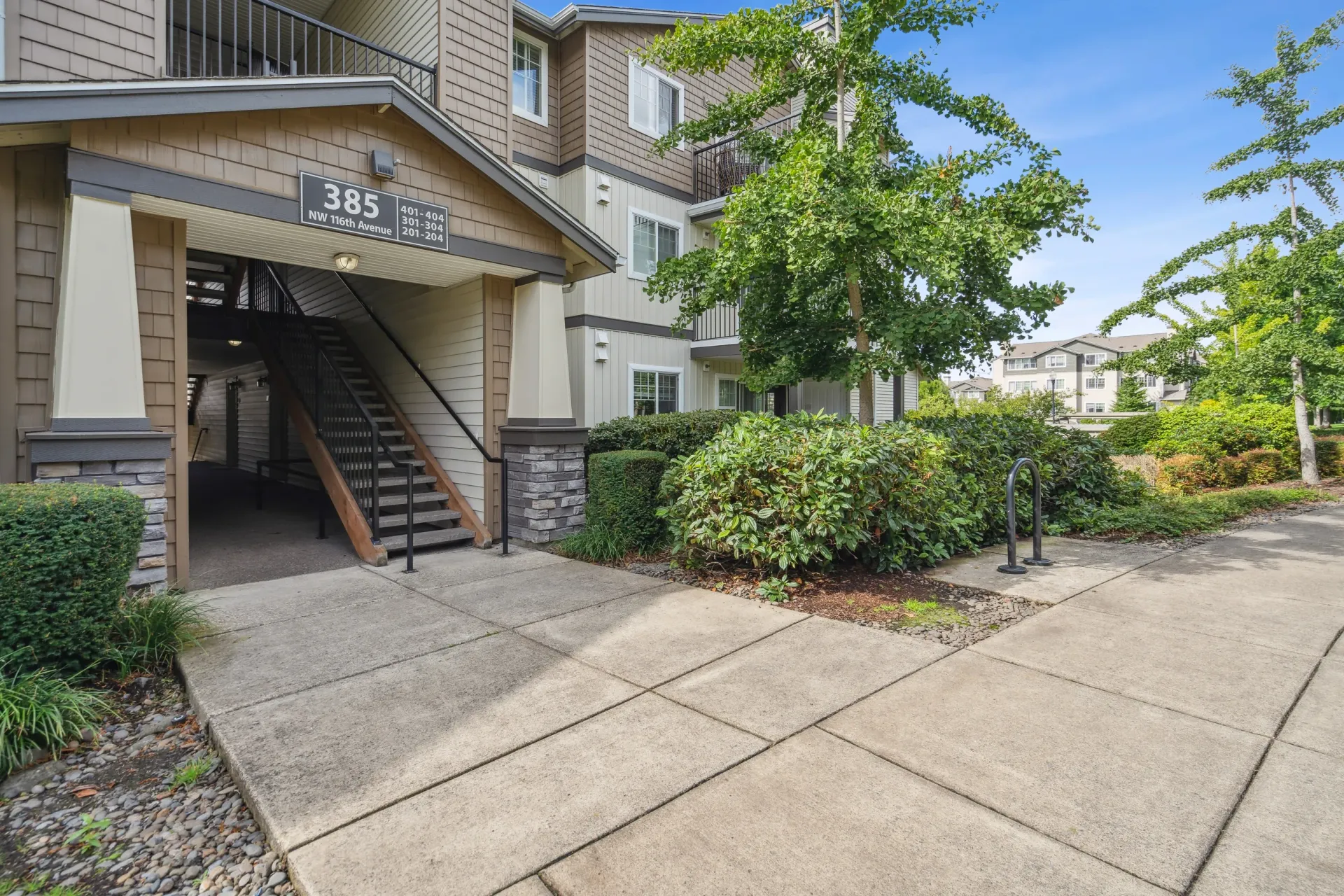 Exterior view of an apartment building entrance with stairs, sign, and landscaping.