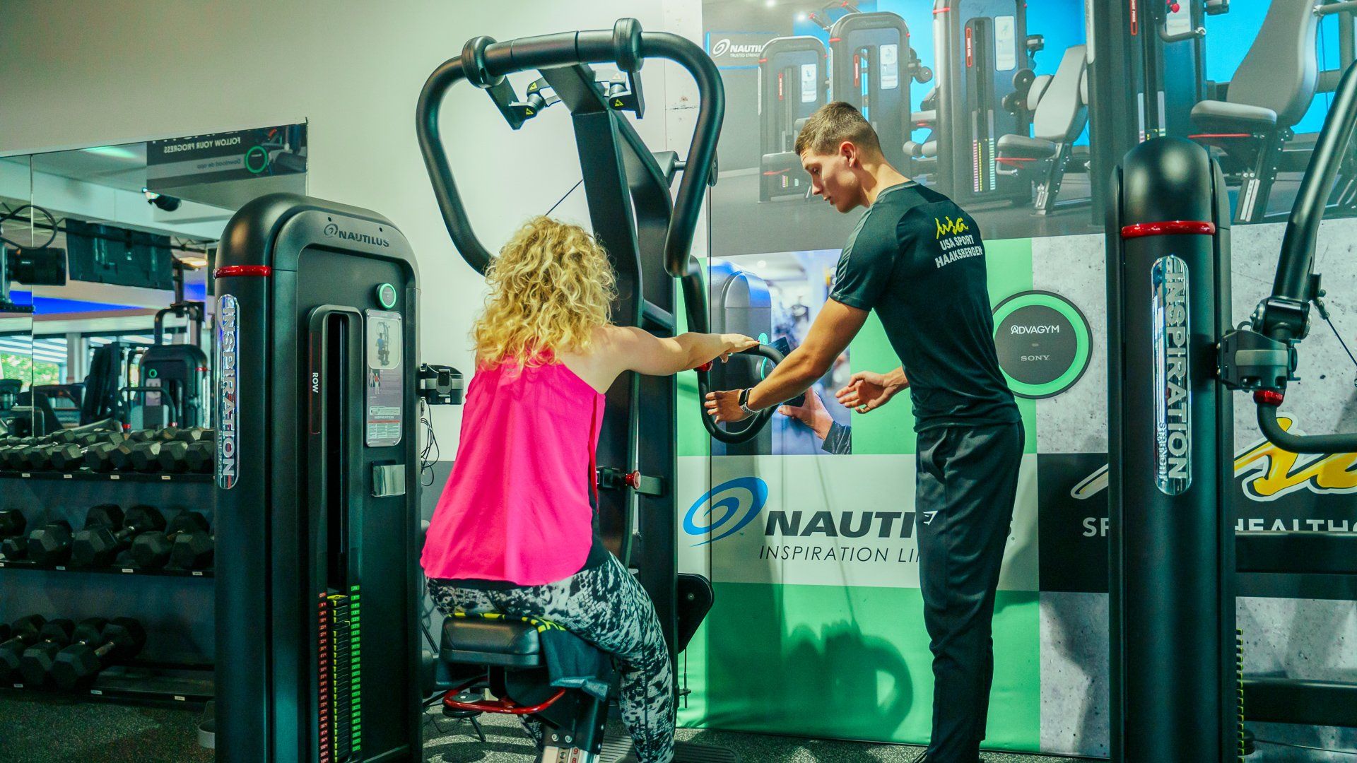 Een personal trainer helpt een vrouw bij een oefeninig op een machine in een sportschool.