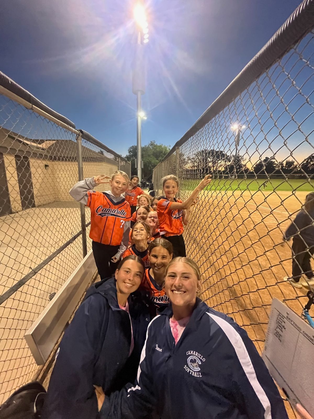 A group of softball players in orange jerseys and navy jackets stand together near a chain-link fence at a sports field.