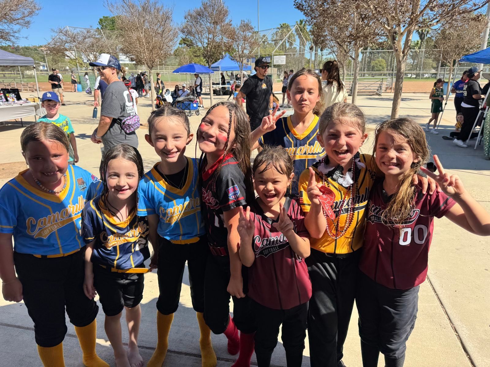 Group of smiling softball players in jerseys, posing outdoors with peace signs.