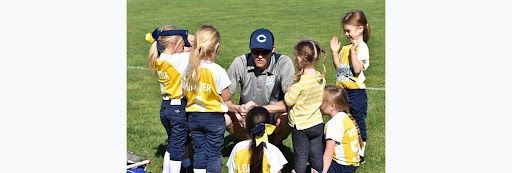 A coach surrounded by young girls, huddled together on a grassy field, likely at a softball game.