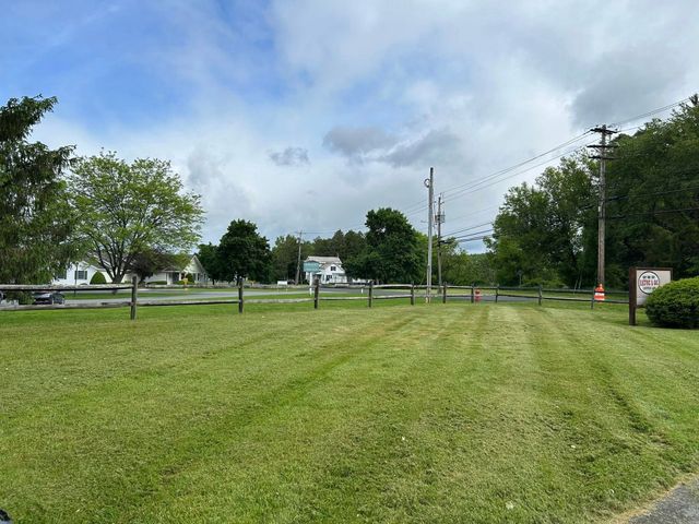 Grassy park field with trees, fence, and cloudy sky; small buildings in the distance.