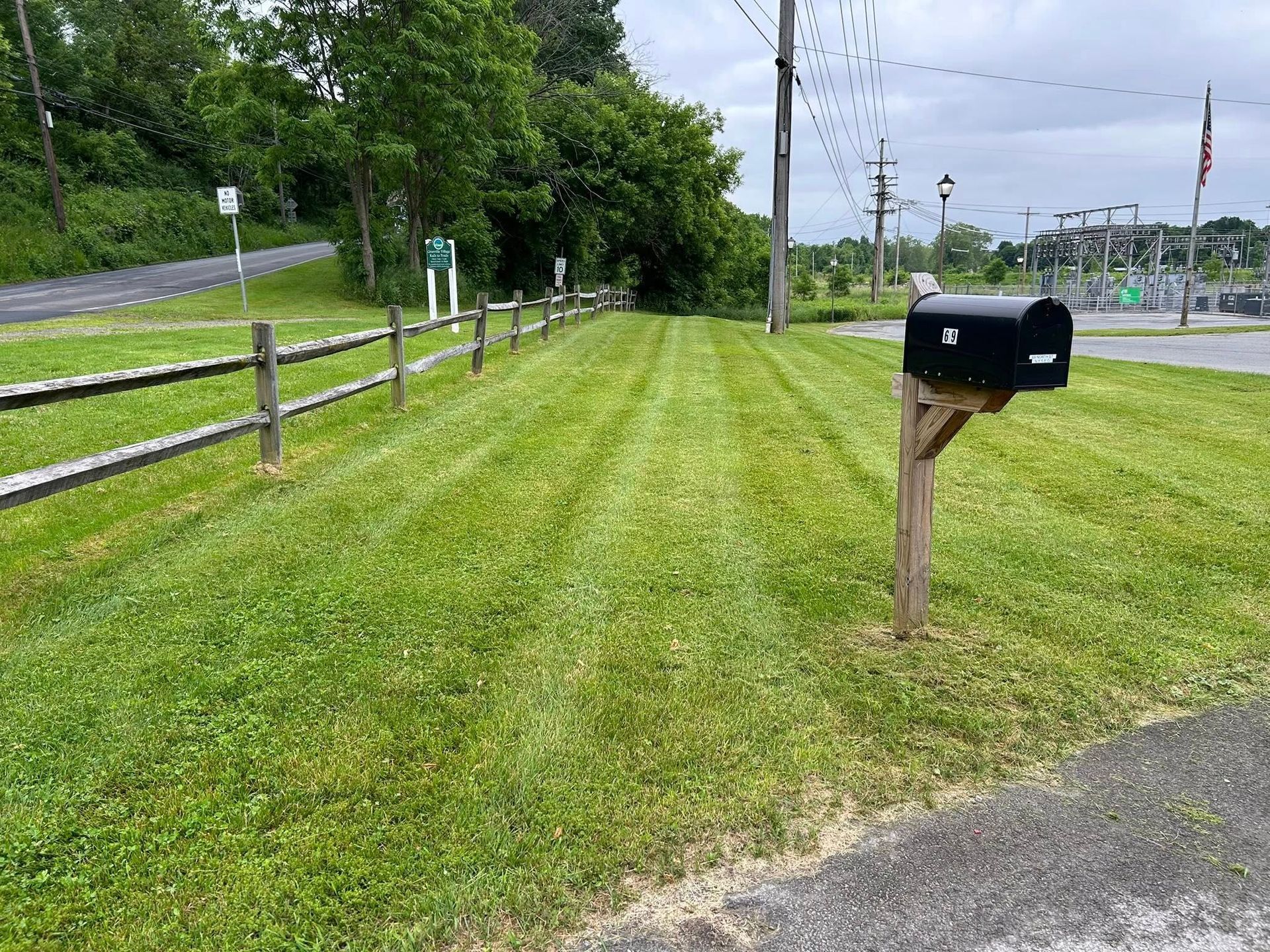 Rural roadside with a black mailbox on a wooden post beside a mowed grassy verge and fence.
