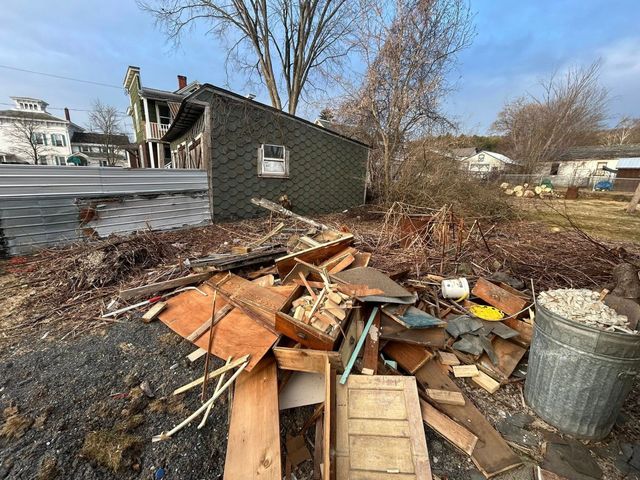 Pile of broken wood and debris in a yard beside a small gray shed on a sunny day.