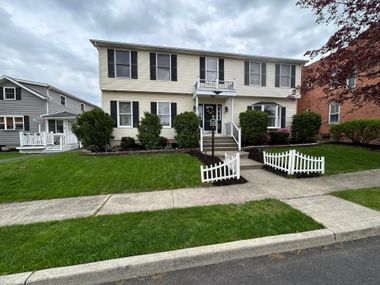 Two-story cream house with black shutters, front lawn, white porch, and a small white fence by the sidewalk