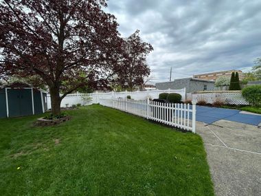 Grassy yard with a white picket fence, large tree, and cloudy sky beside a street.