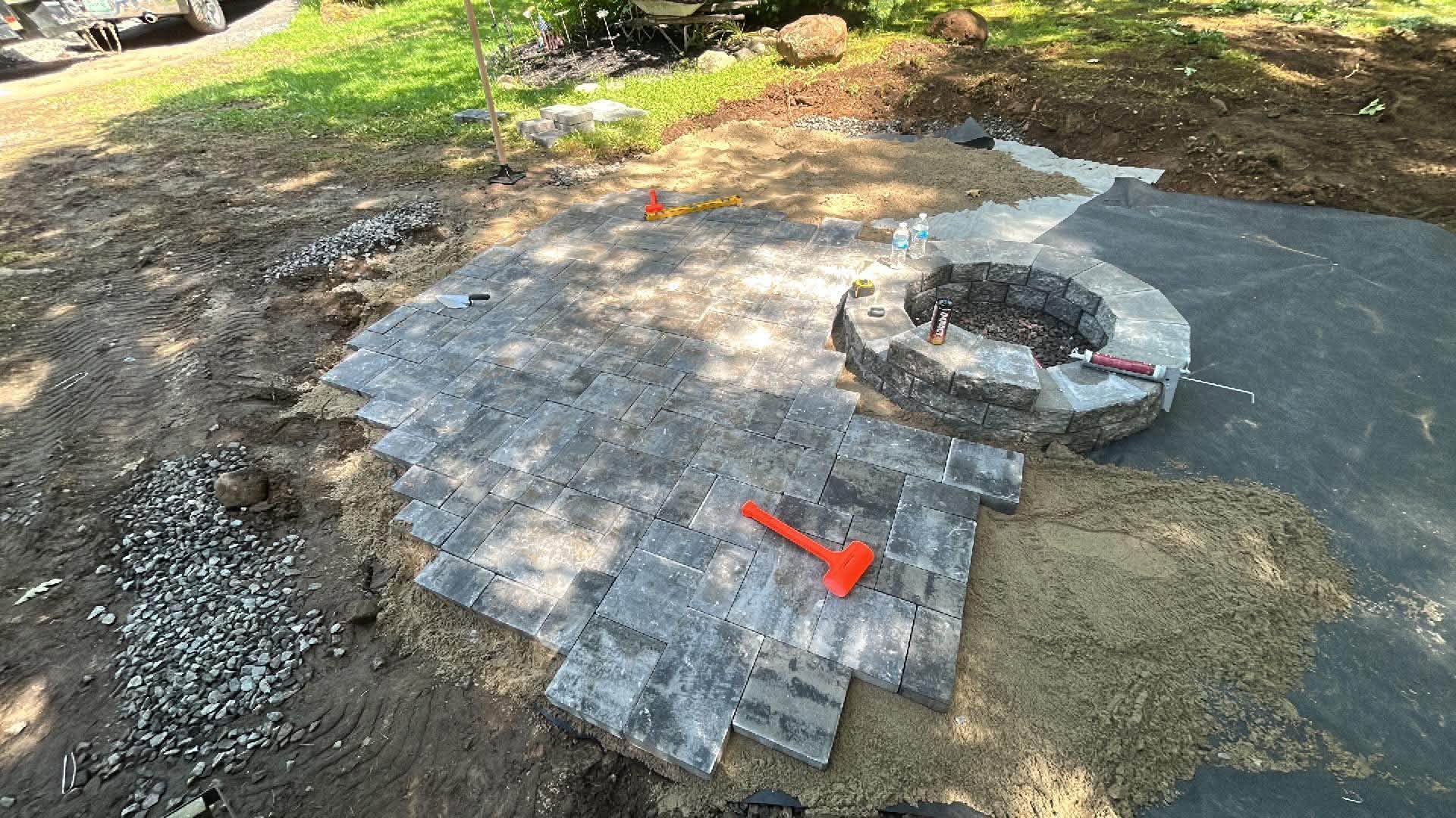 Stone patio under construction with gray pavers and a circular fire pit in a yard