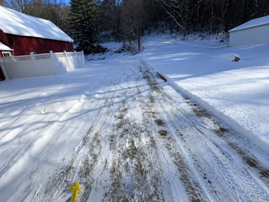 Snow-covered driveway with tire tracks beside a red building and trees in winter