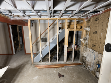 Partially demolished basement with exposed studs, stairway, and debris on the floor