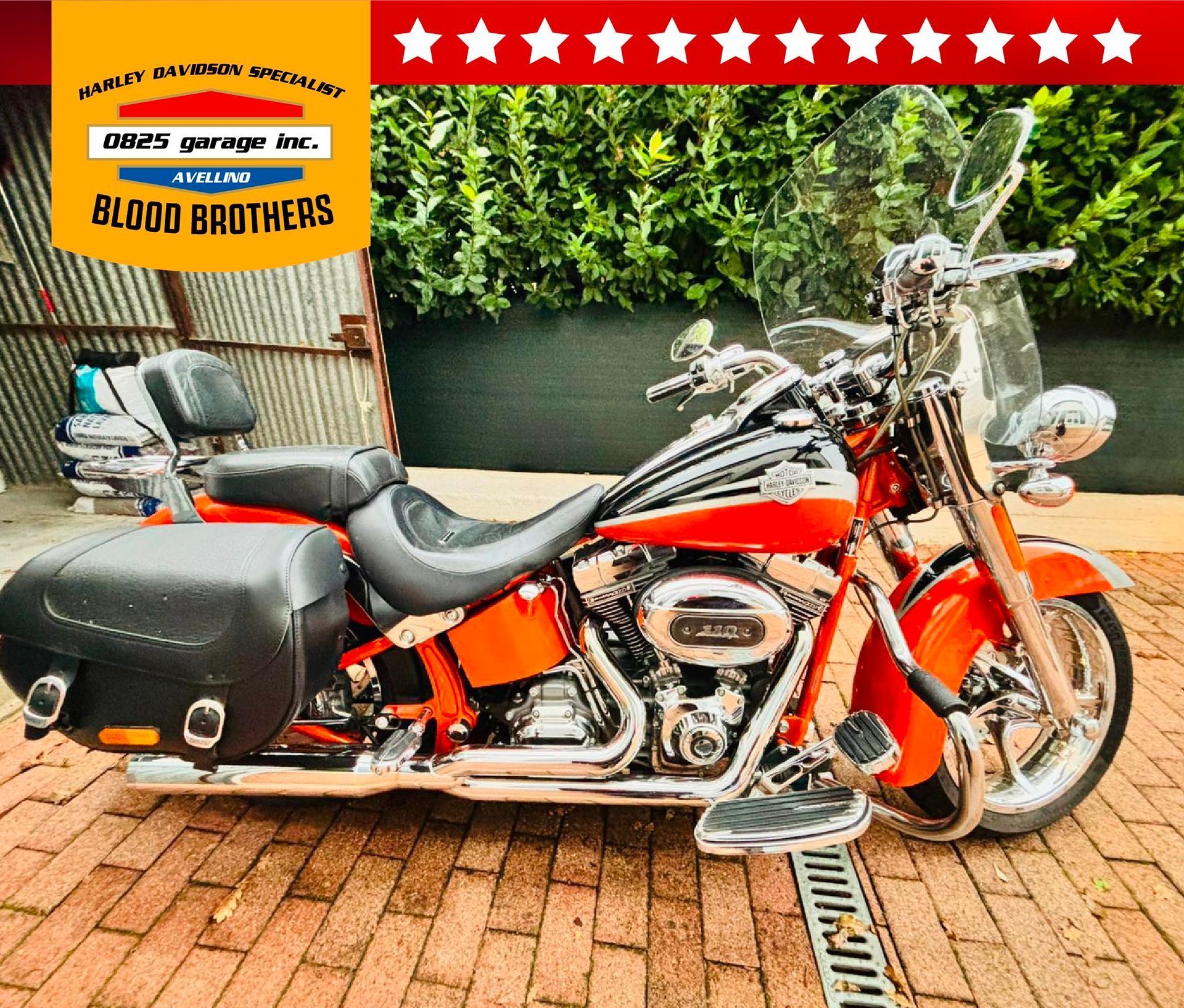 Una motocicletta Harley-Davidson rossa con borse laterali nere parcheggiata su una superficie di mattoni, accanto a un muro di lamiera ondulata.