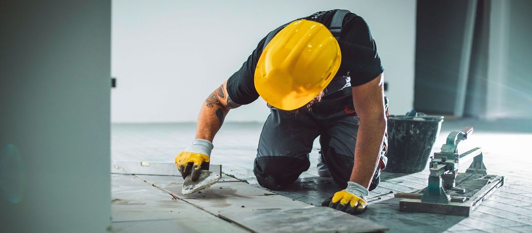 Un operaio edile che indossa un casco e dei guanti è inginocchiato sul pavimento.
