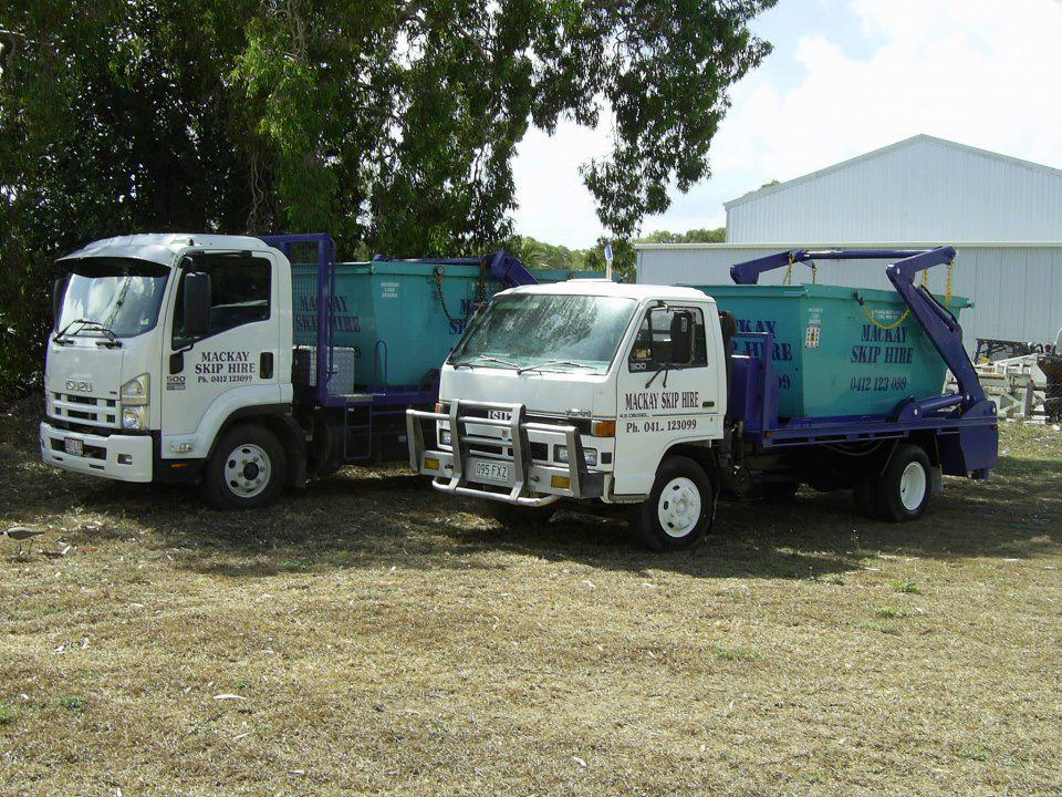 Two White and Blue Roll-off Trucks With Waste Containers Parked on Dry Grass — Mackay Skip Hire In Andergrove, QLD