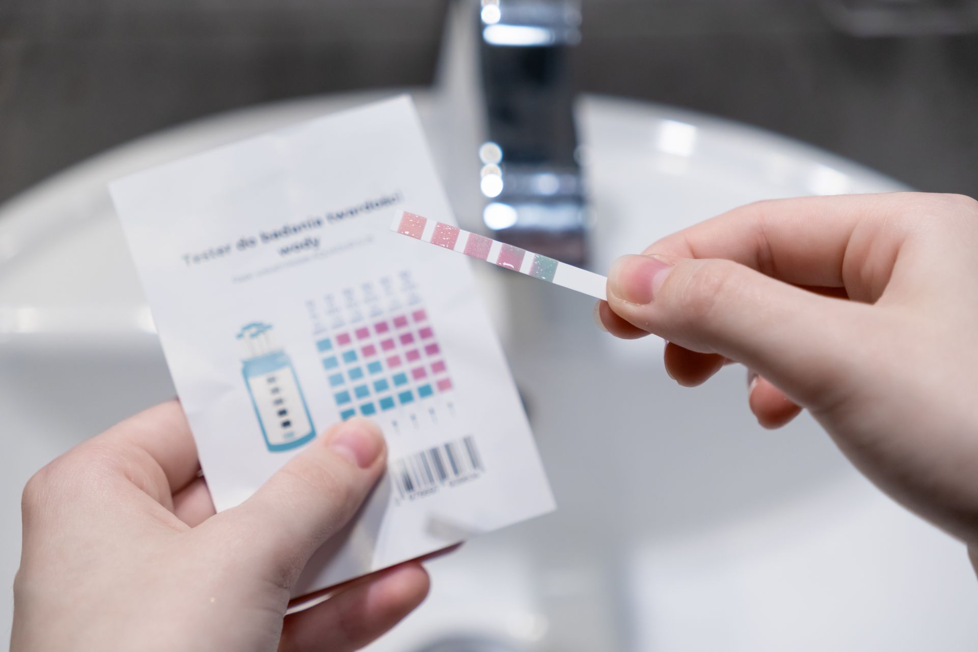 Hands holding a chemical test strip against its color-coded reference card, with a sink faucet in the background.