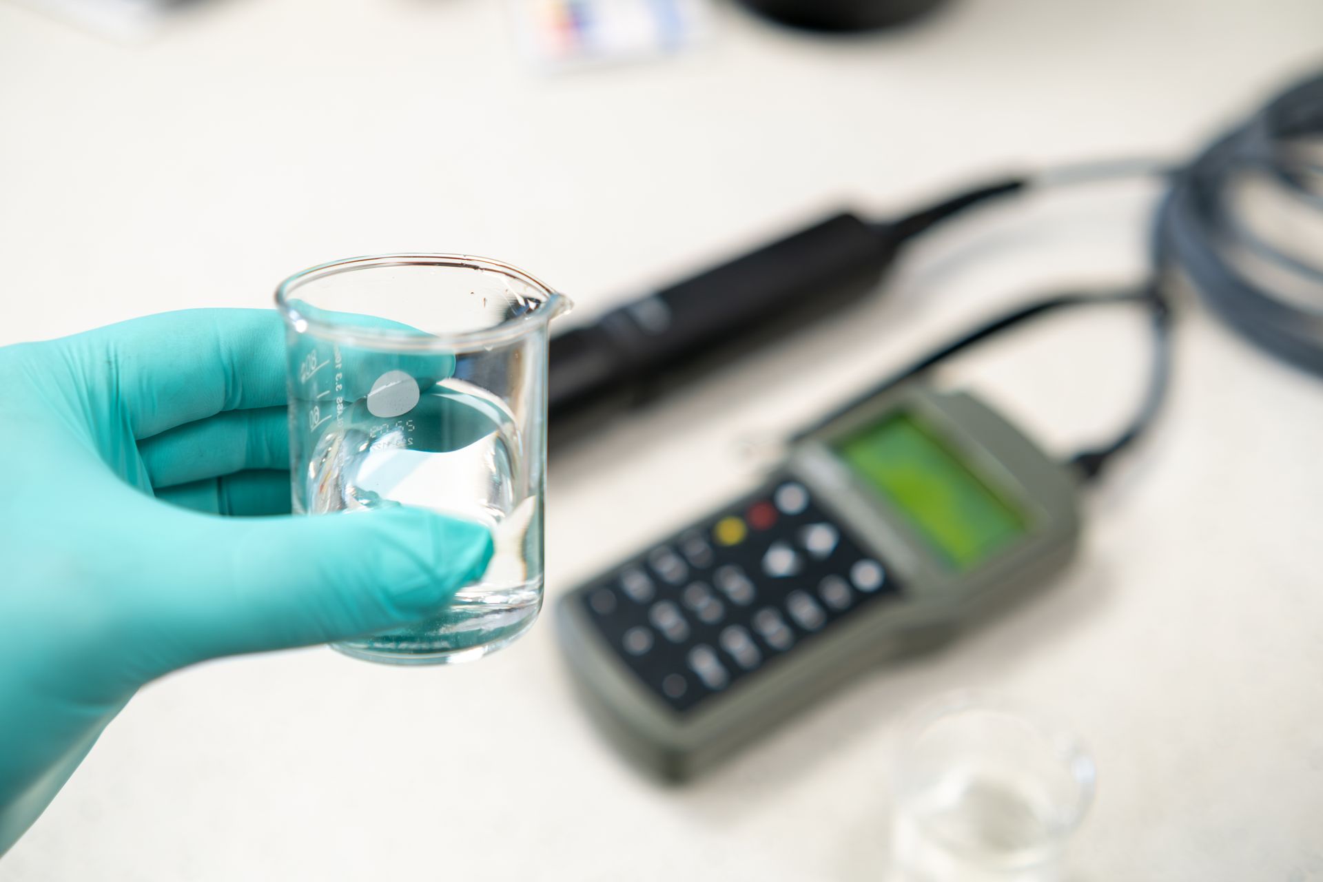 A gloved hand holds a beaker of clear liquid near a scientific water quality meter in a laboratory setting.