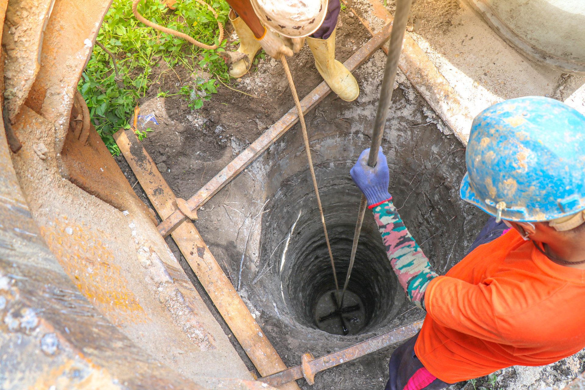 A worker in an orange shirt and blue hard hat uses a metal tool to guide a drill into a deep, circular hole in the ground.