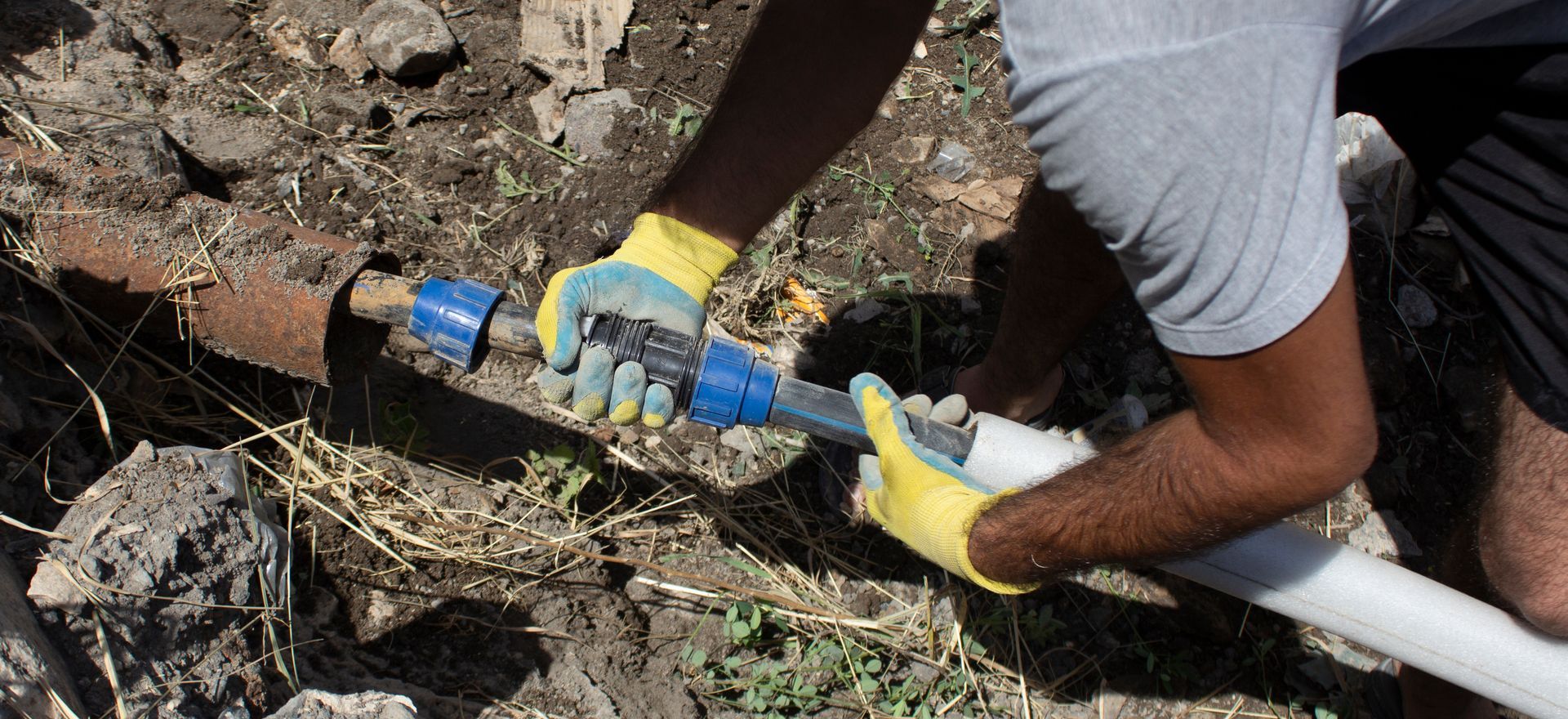 A person wearing yellow gloves connects a new white pipe to an existing dark pipe in the ground using a blue coupling.