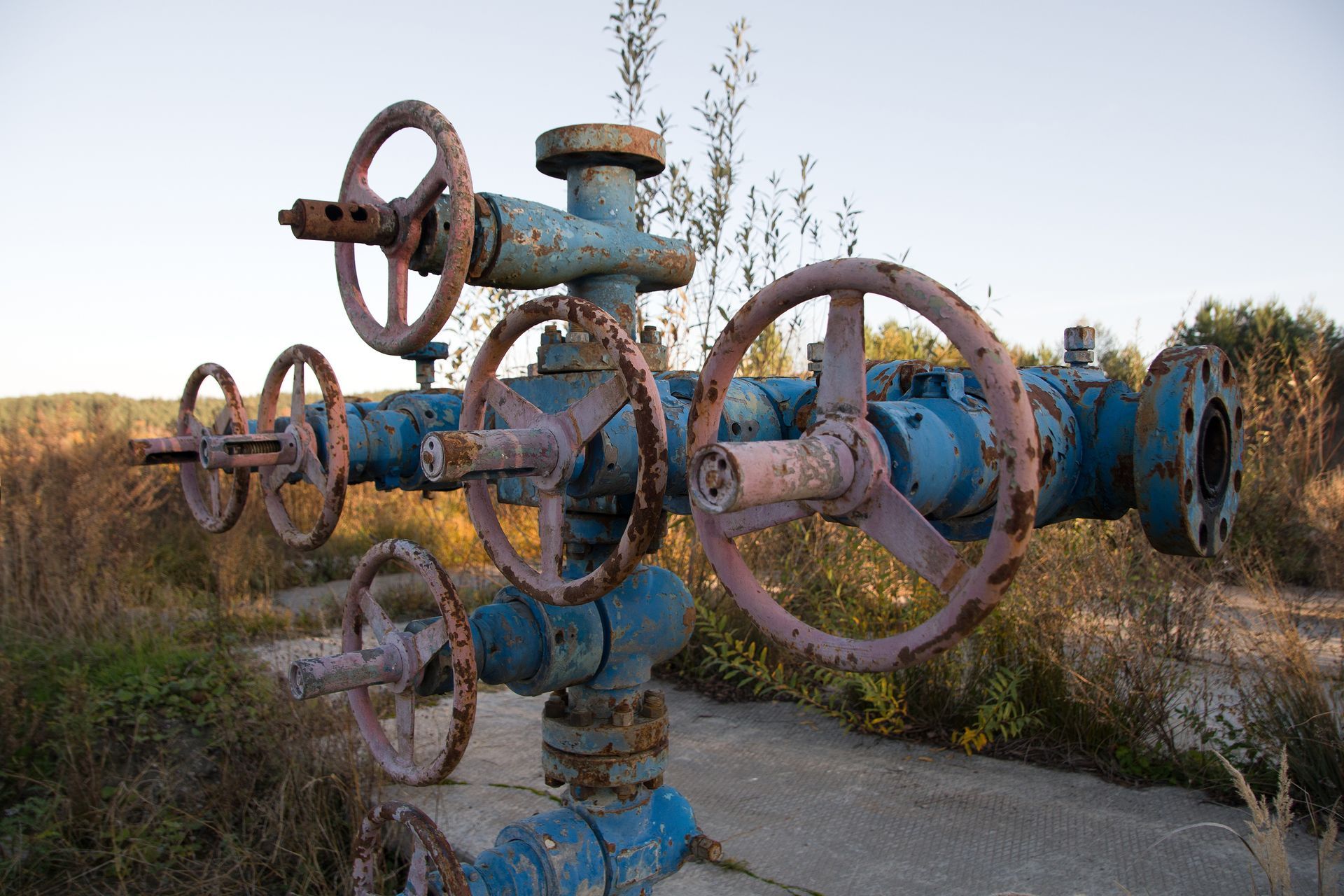 A weathered, blue industrial wellhead with rusted handwheels situated outdoors in a field of dry vegetation.