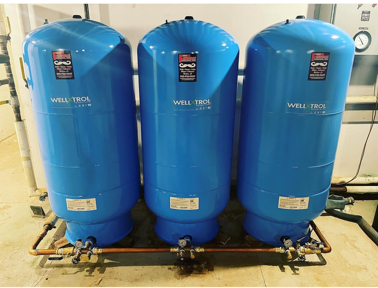 A person's hand touches a white PVC pipe connected to a blue water pressure tank and a cylindrical water filter.