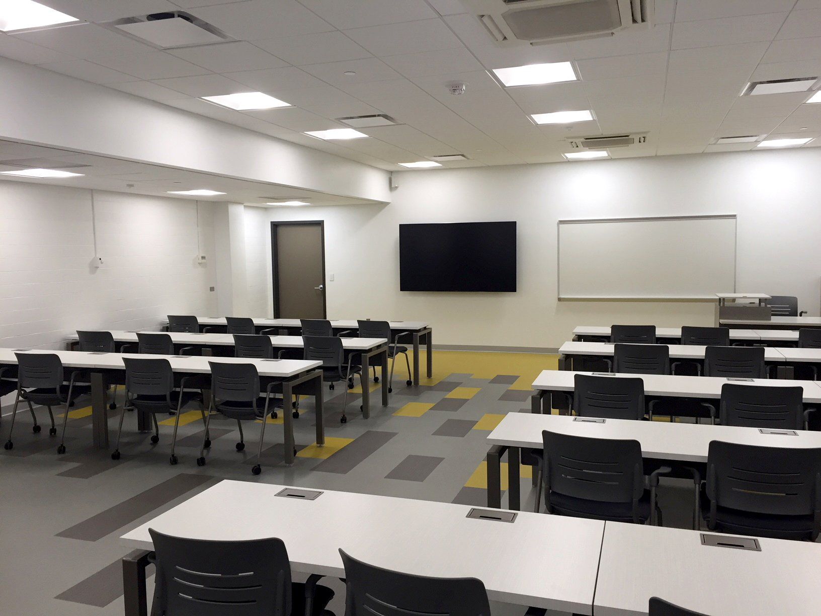 A large classroom with tables and chairs and a large screen on the wall.