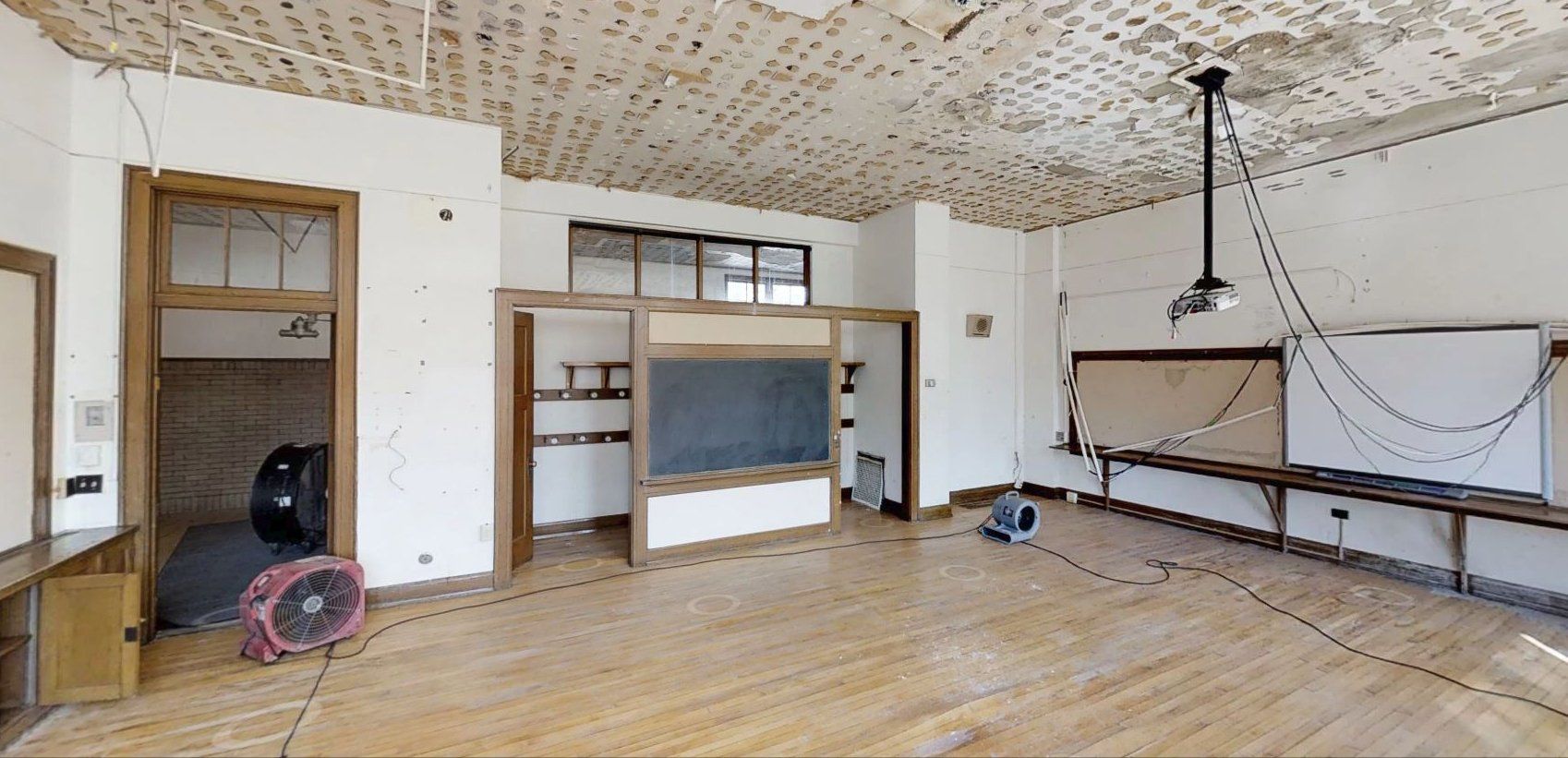 An empty classroom with a blackboard and a fan on the floor.