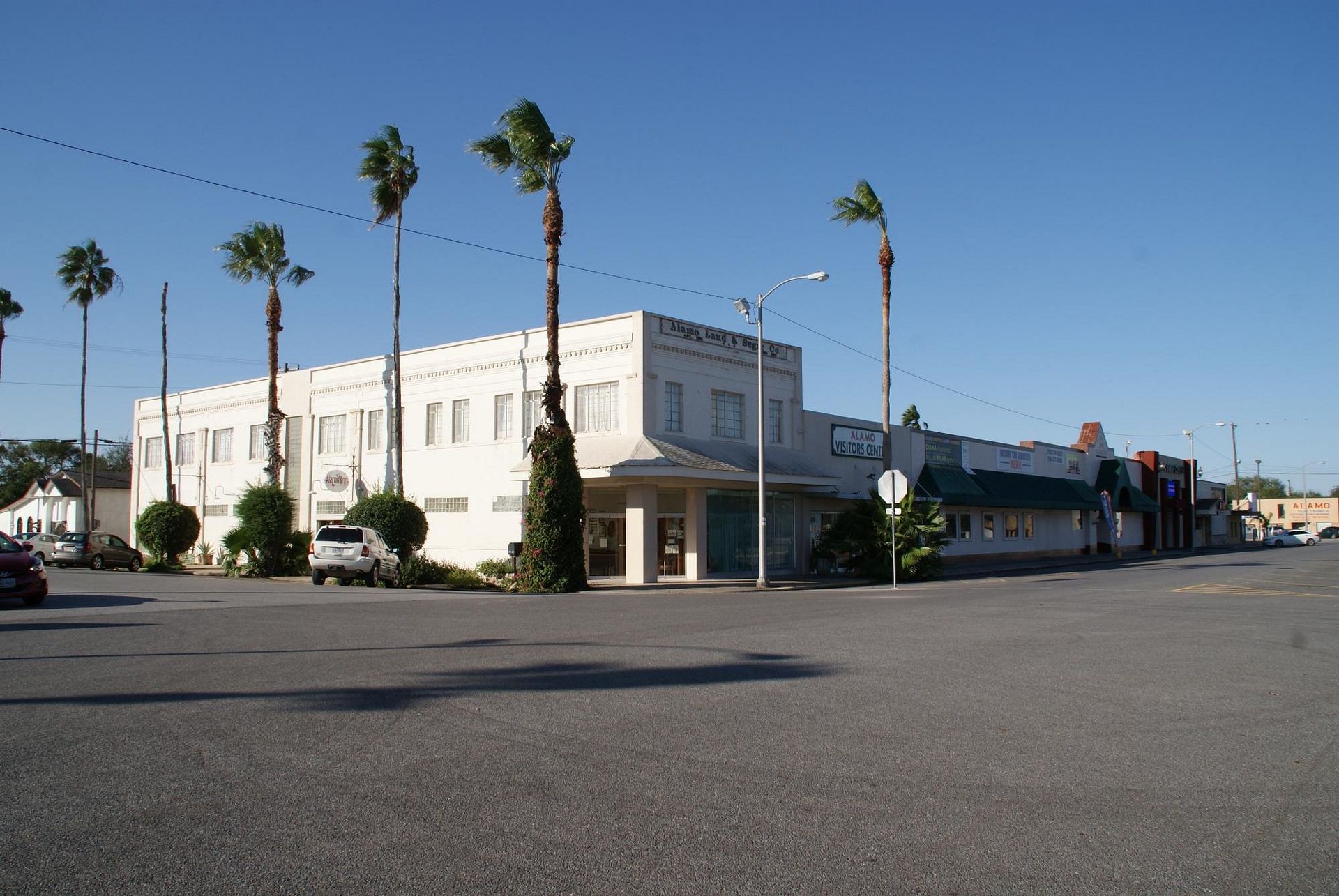 A multi-story beige stucco building with palm trees on a paved lot under a clear blue sky.