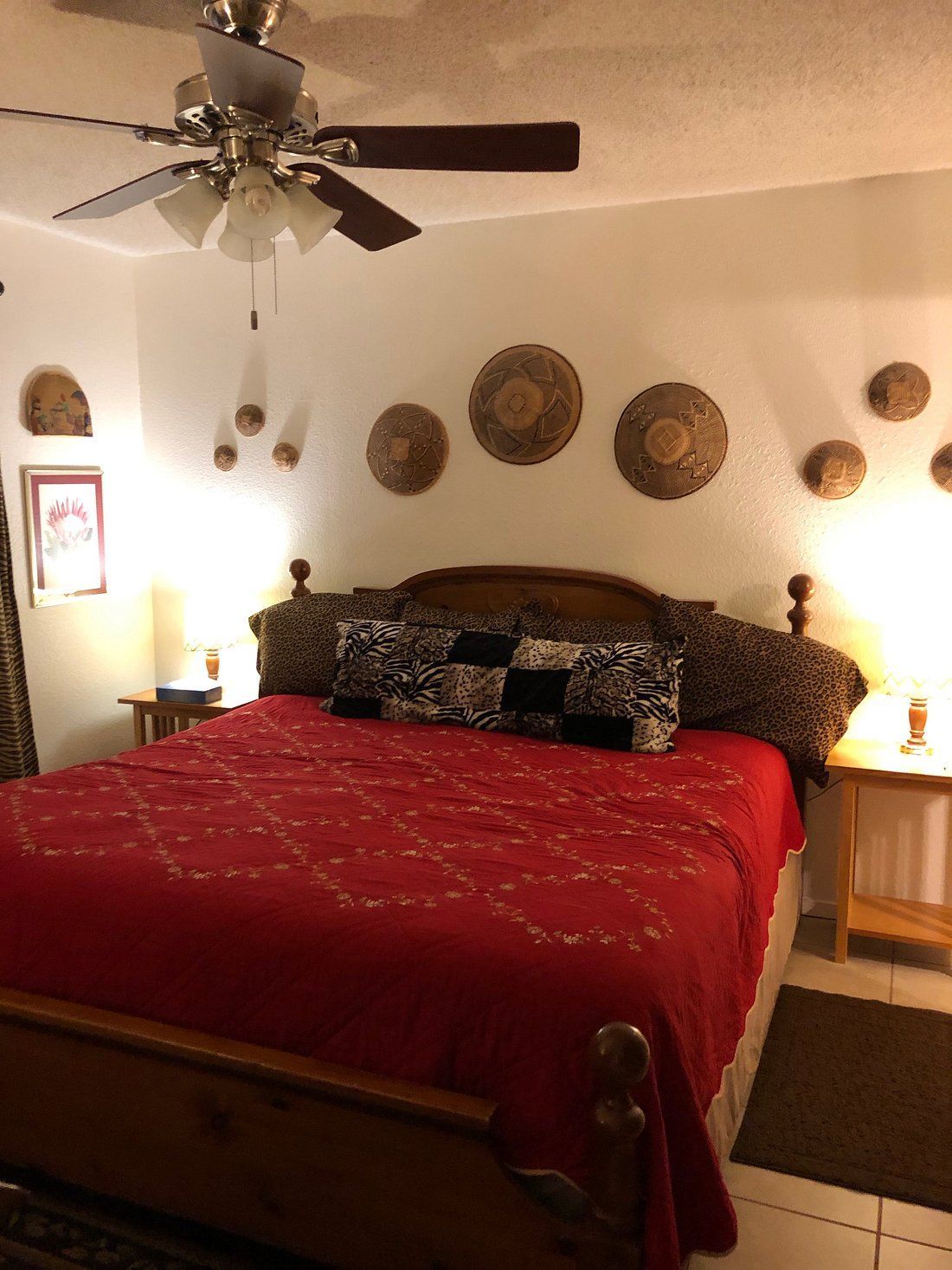 A bedroom with a red bedspread on a wooden bed frame, decorated with wall-mounted straw hats, under a ceiling fan.