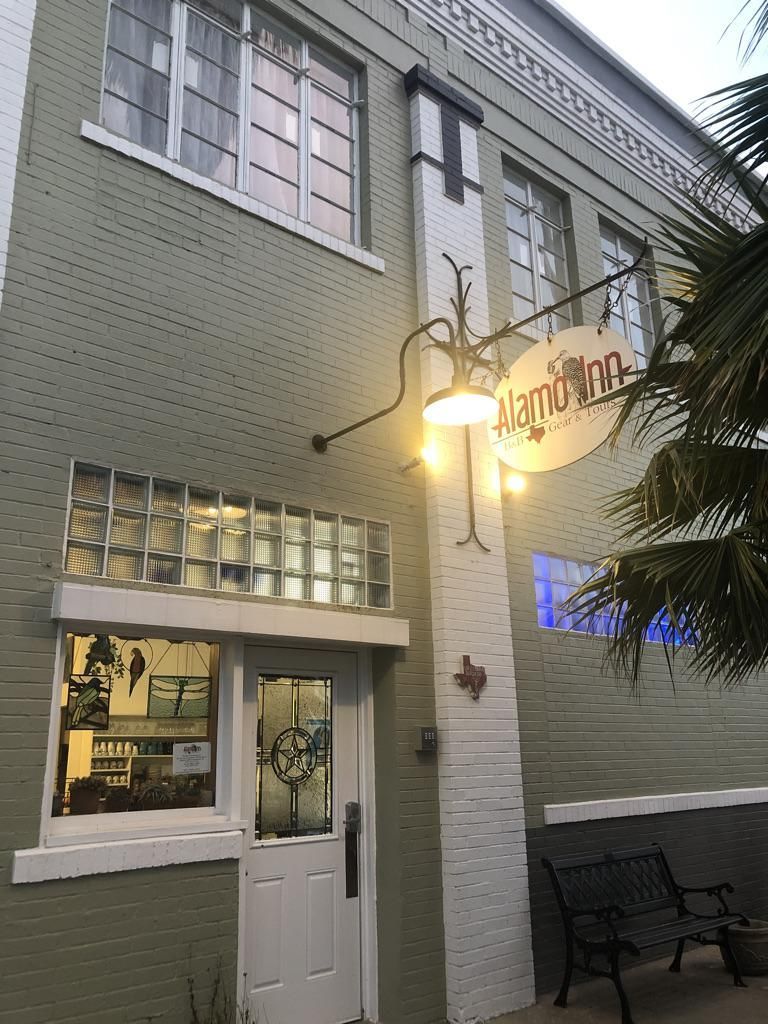 A green brick building exterior features a white door, glass block windows, and a round sign hanging from a metal light.
