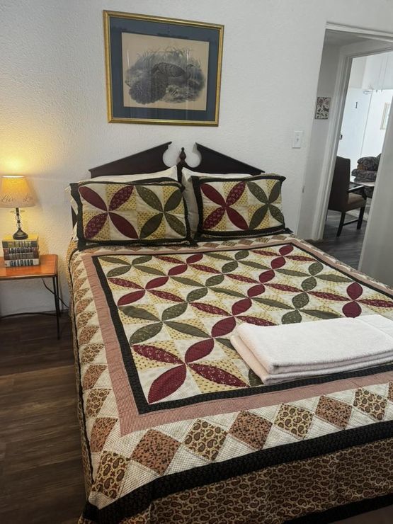 A bed with a quilt featuring a geometric floral pattern in red, green, and tan, with a folded white towel on top.