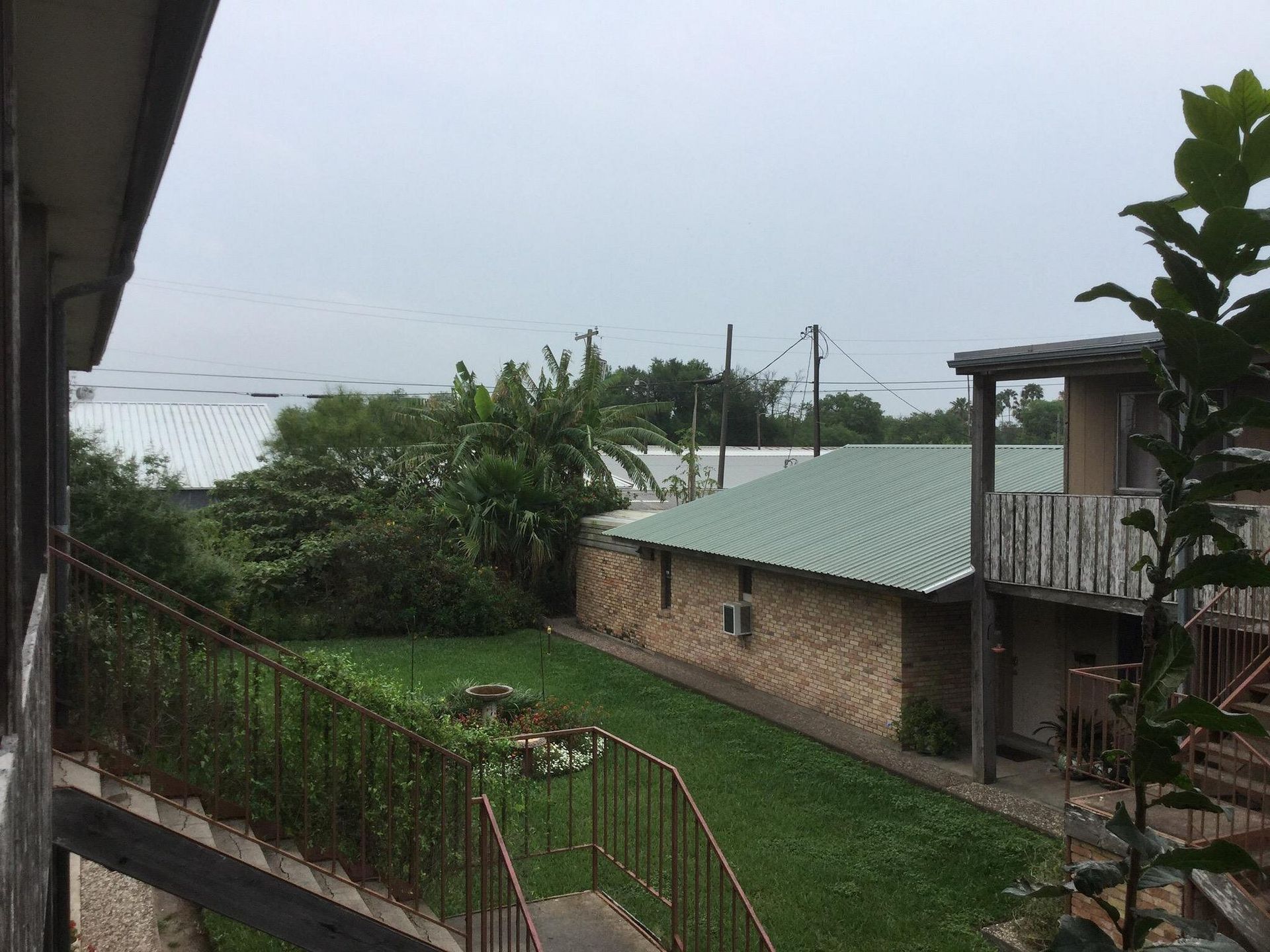 A view of a grassy courtyard between brick buildings with a metal staircase, lush green trees, and an overcast sky.