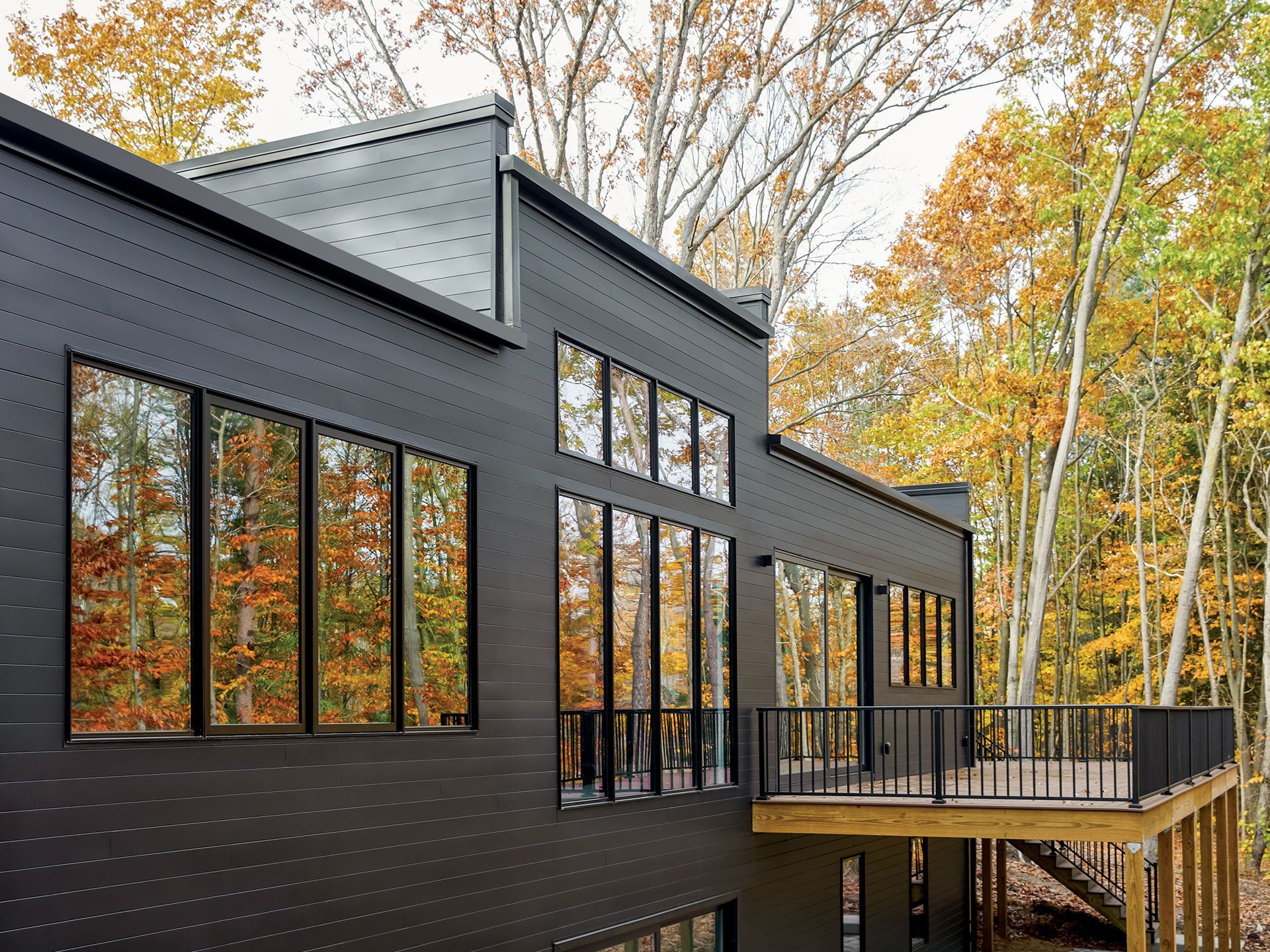 Modern black house with large windows reflecting autumn trees, and a deck.