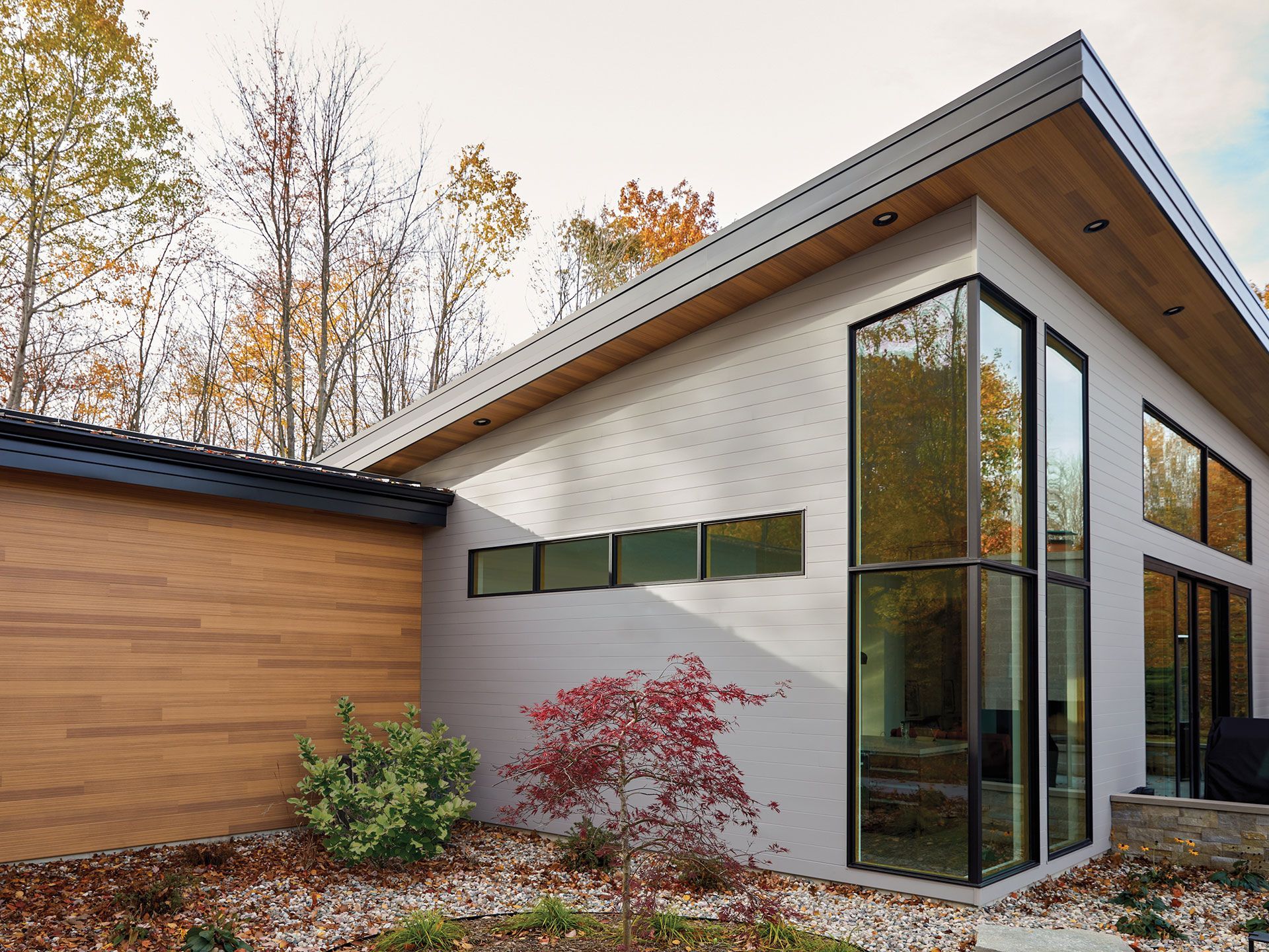 Modern white house with large windows, wood siding, and autumn foliage in the background.