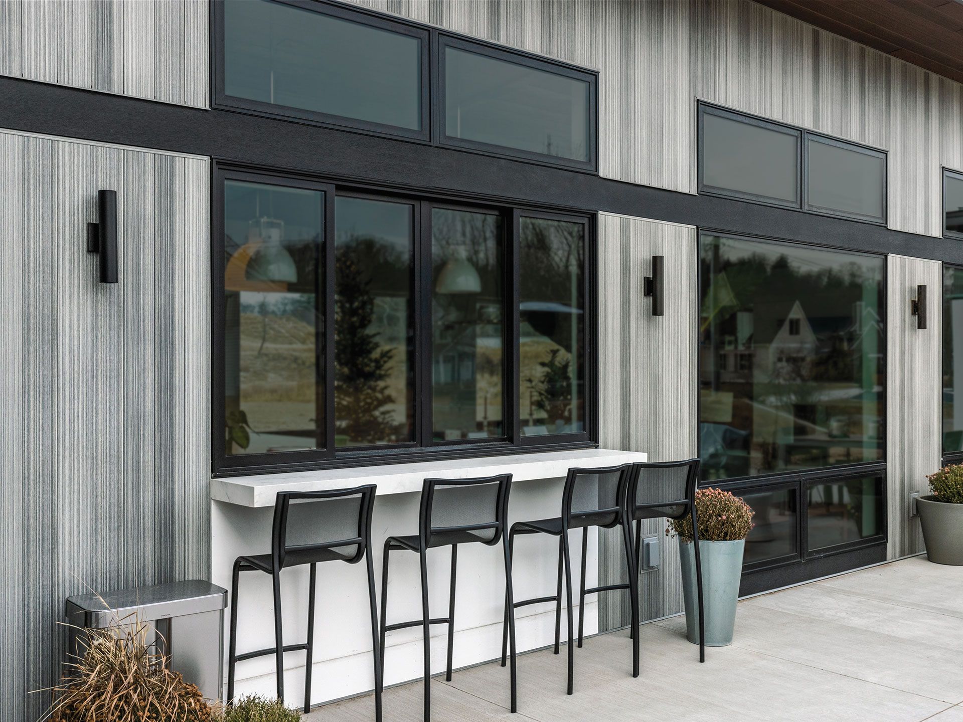 Exterior of a modern building with bar seating. Gray siding, black trim, and large windows.
