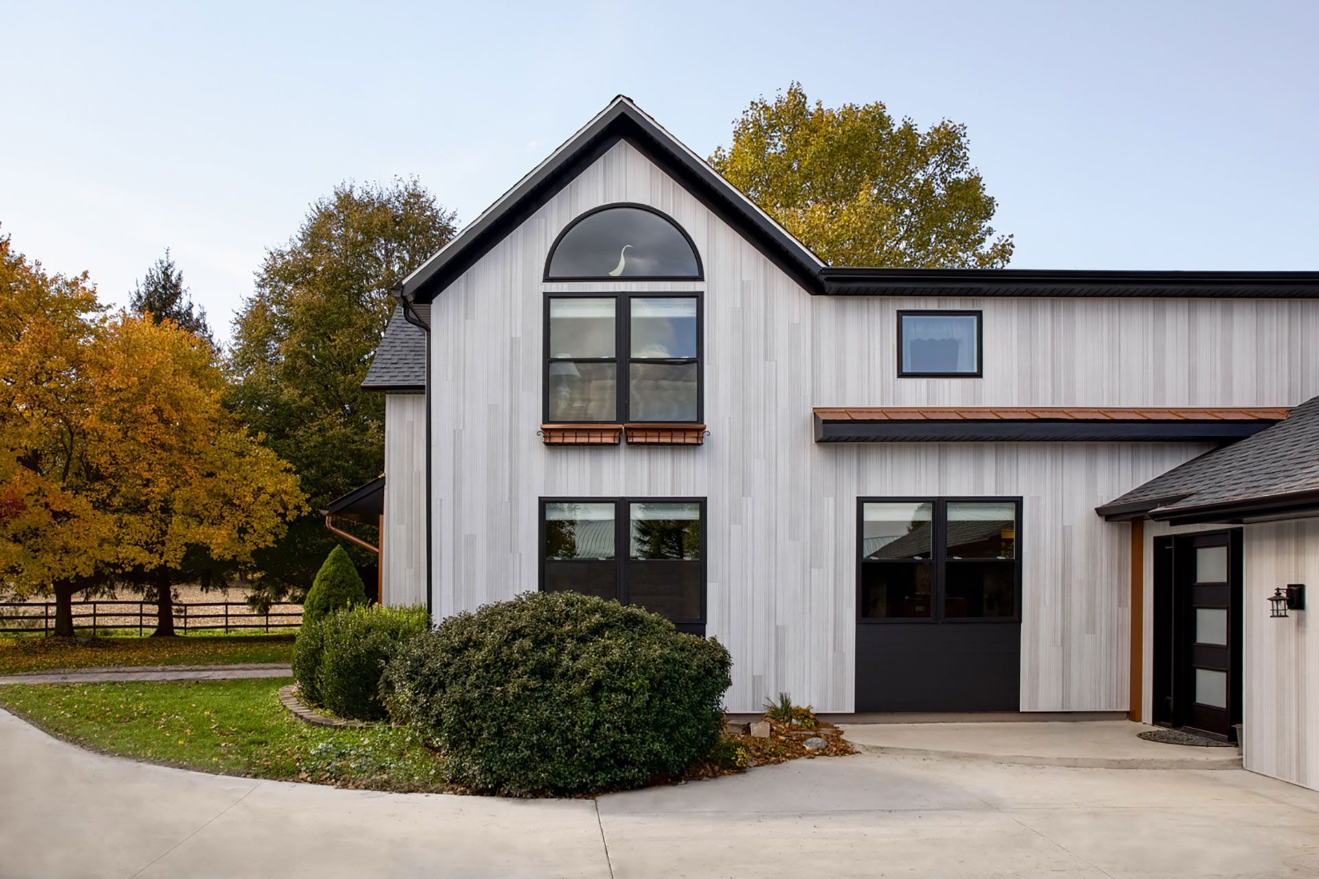 Modern white barn-style house with black trim and windows, surrounded by autumn trees and a driveway.