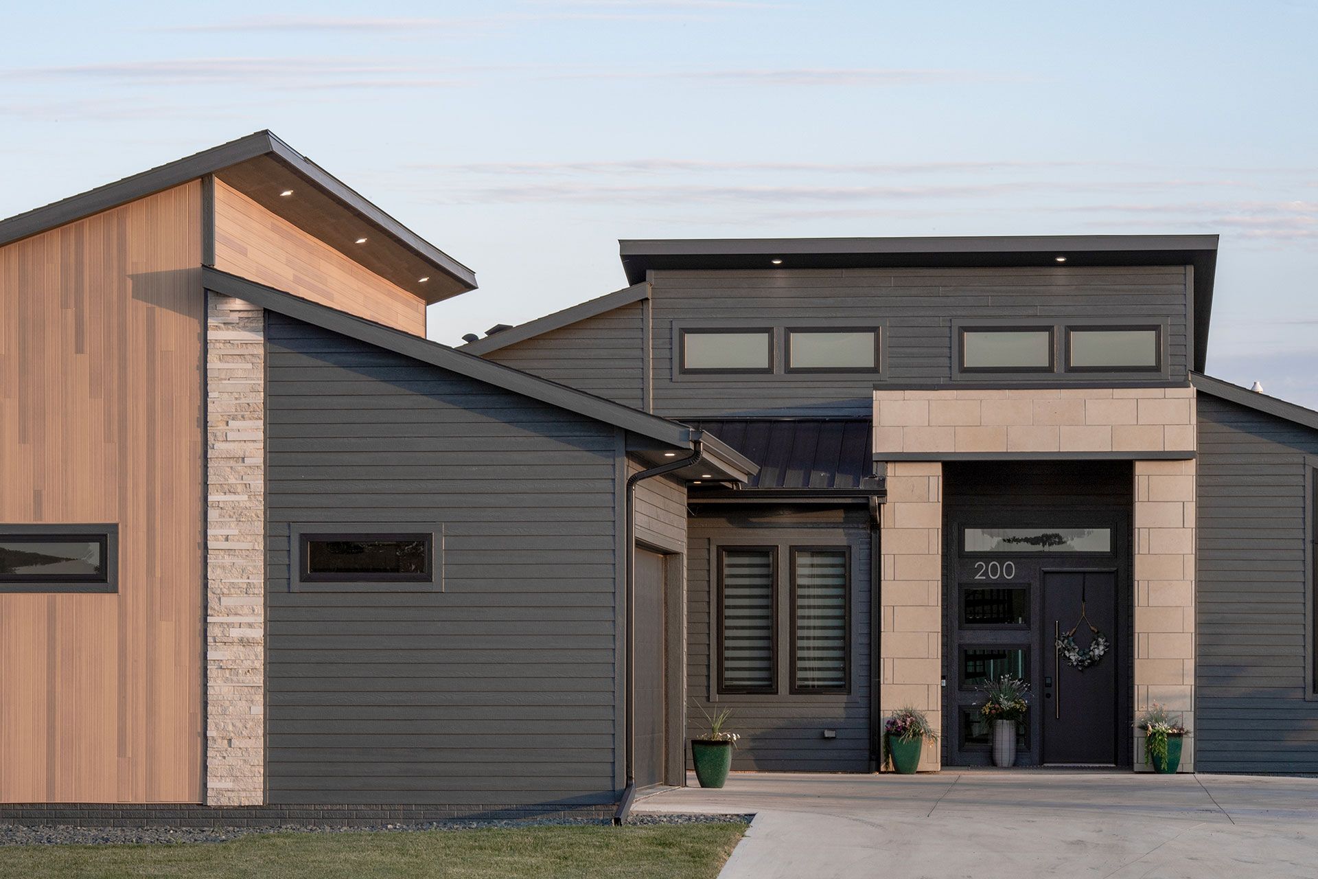 Modern house exterior, grey siding, light stone accents, wood panels, dark door, potted plants.