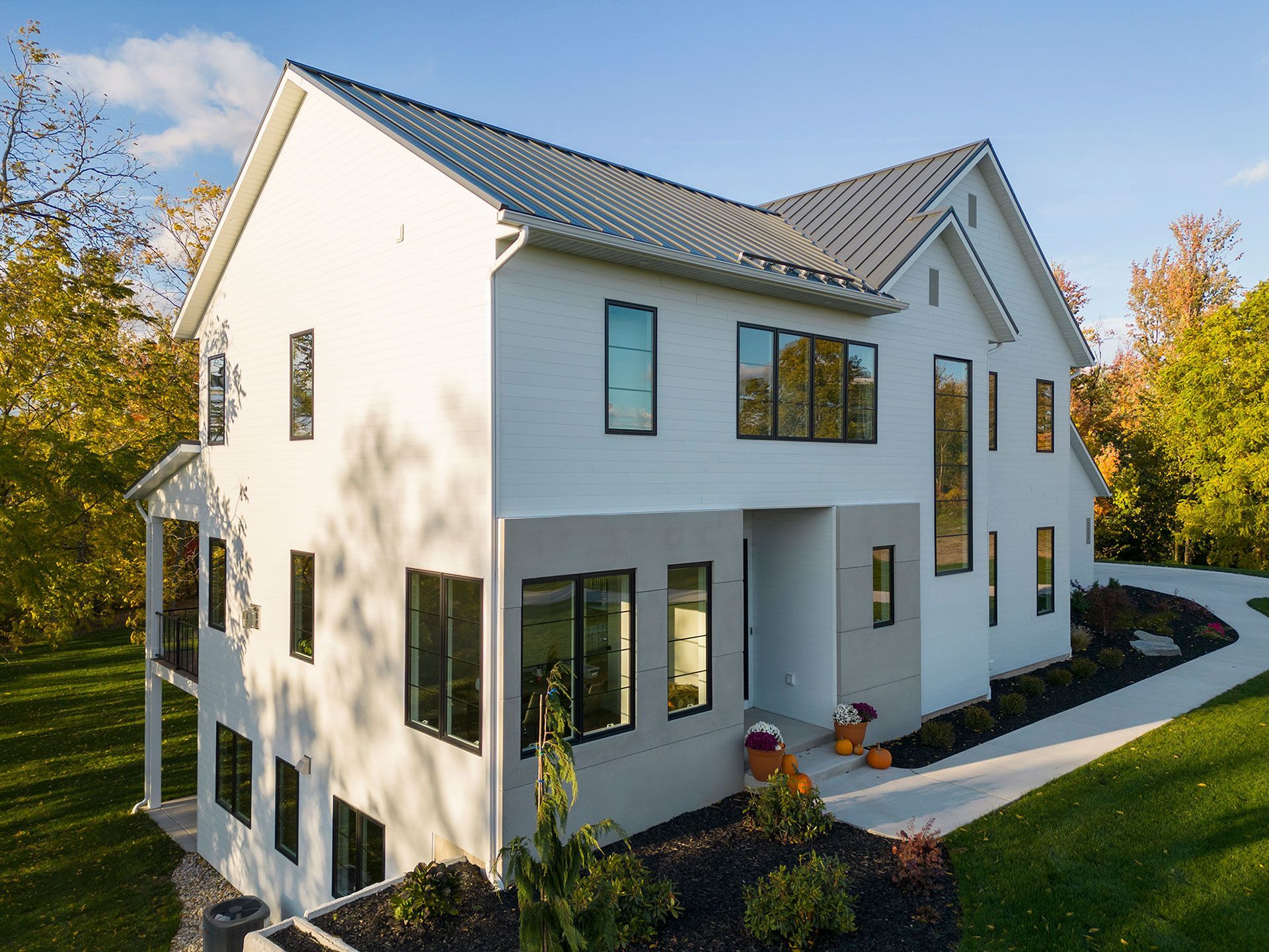 White modern house with black-framed windows, metal roof, and pumpkins on the front path, set in a green, sunny yard.