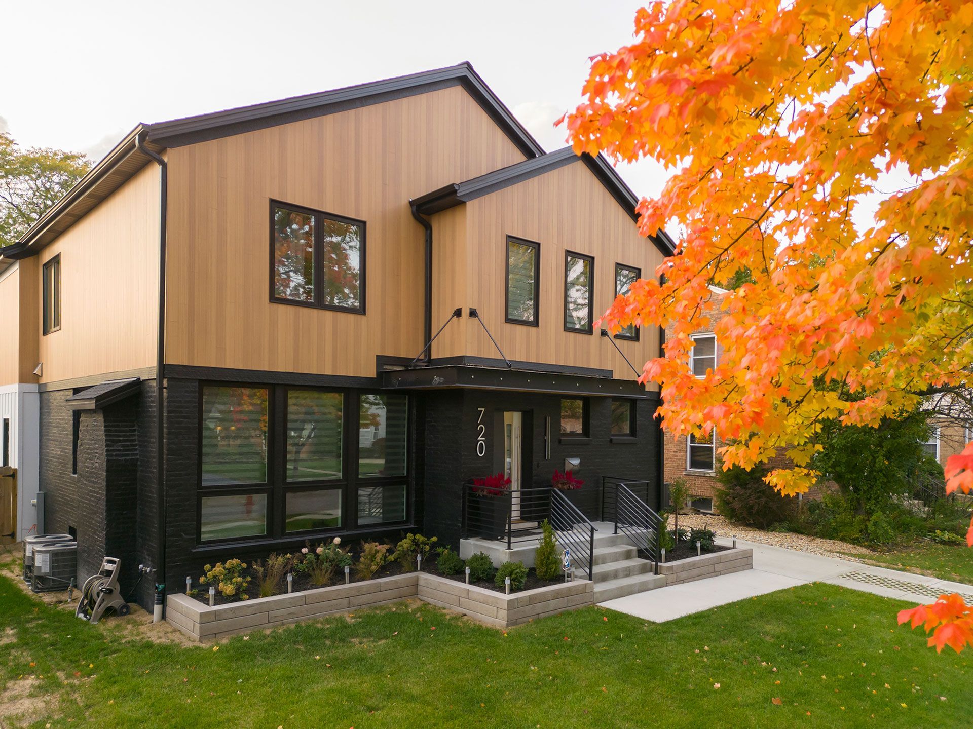 Modern two-story house with black and brown siding, a lush yard, and a colorful fall tree.