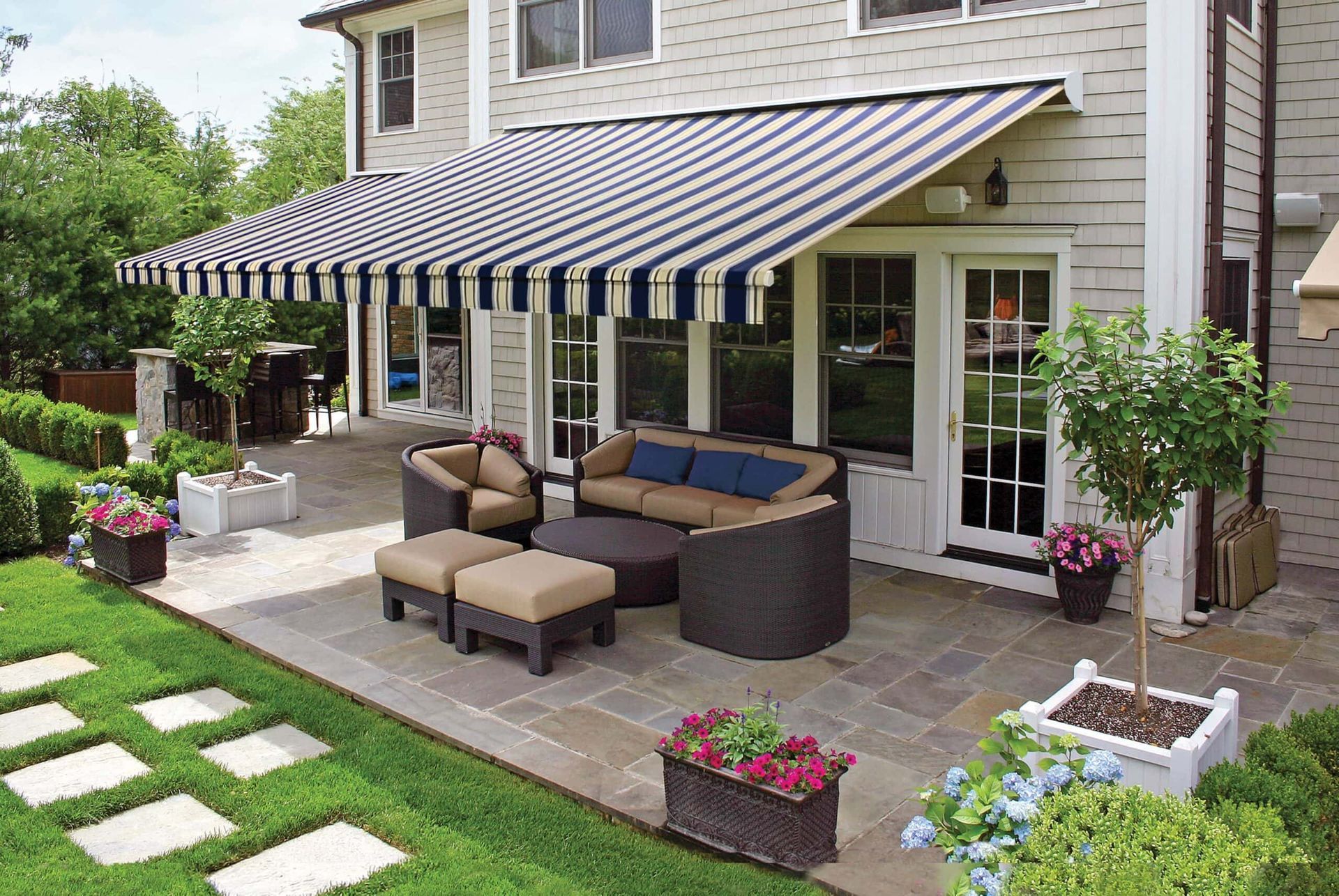 A patio with outdoor furniture, a striped awning, and flower beds, beside a house.