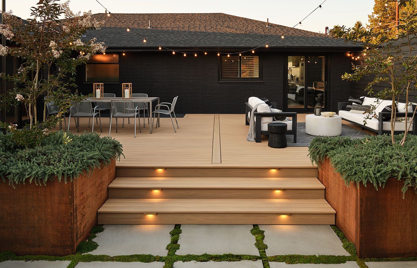 Wooden deck with steps leading to outdoor seating and dining area, black house, lit by string lights.