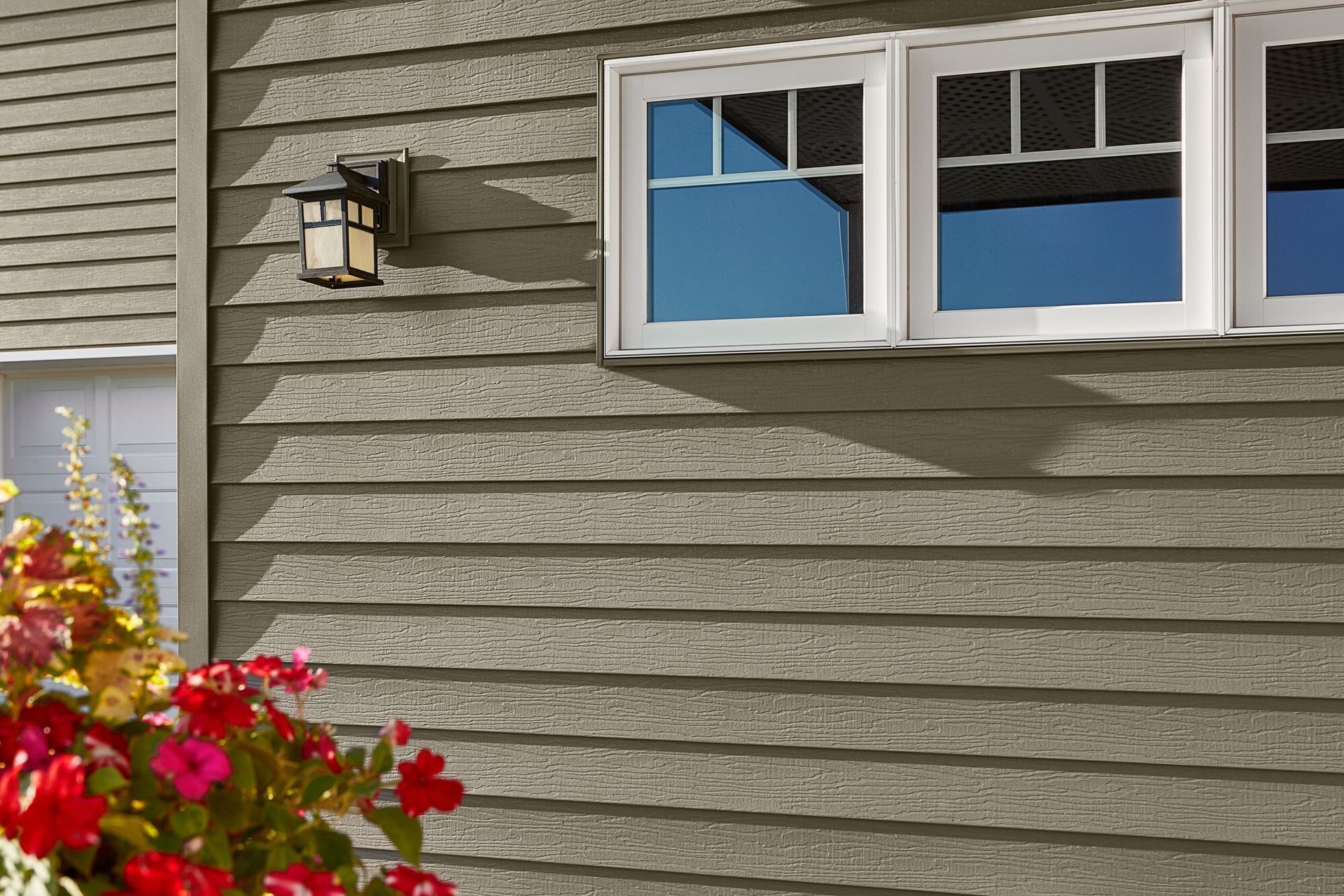 Exterior of a house with gray siding, white window trim, and a hanging light fixture, with red flowers in the foreground.
