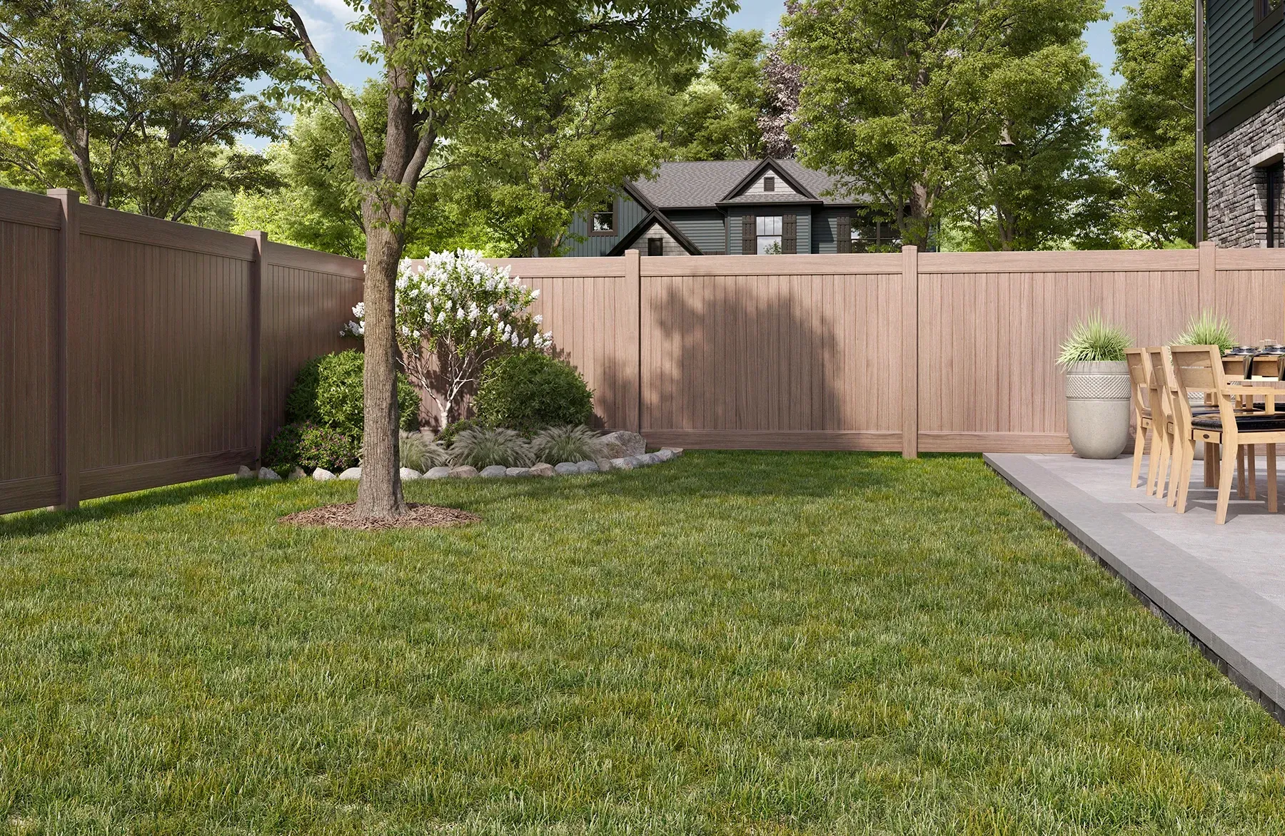 Brown privacy fence surrounds a backyard with green grass, a tree, and a stone patio with a table and chairs.