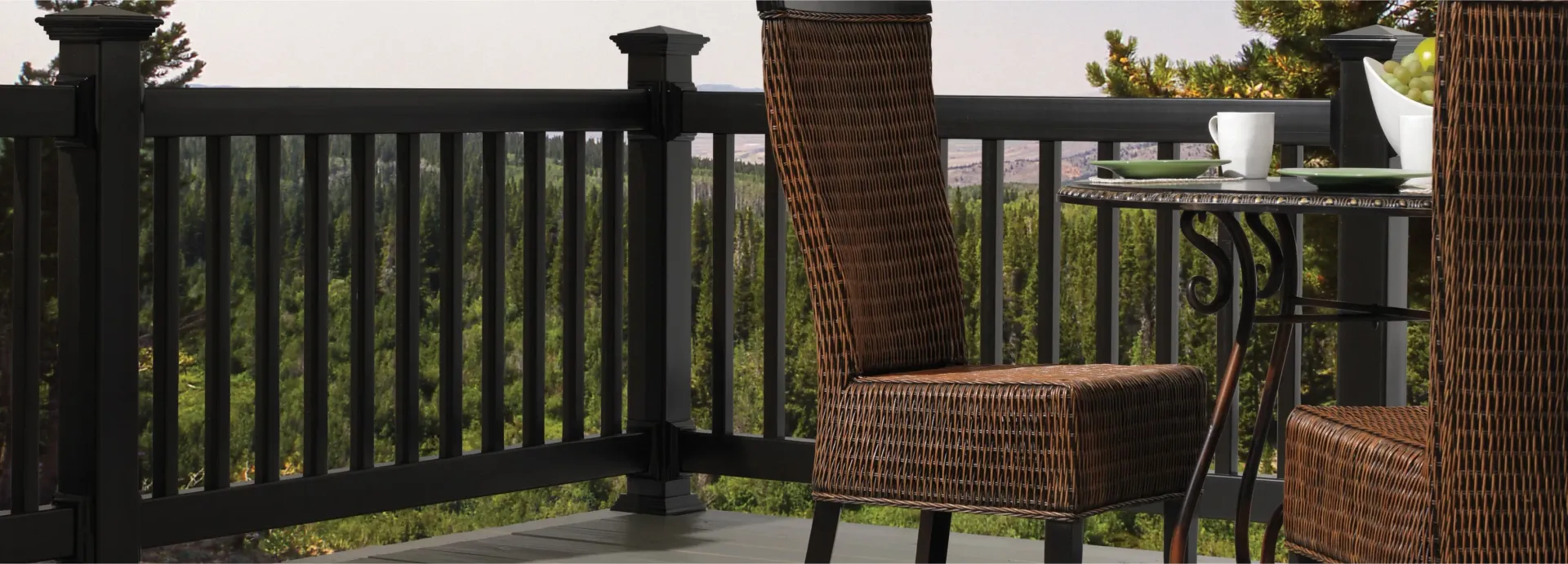 Wicker chairs and table on a balcony with black railing overlooking trees.