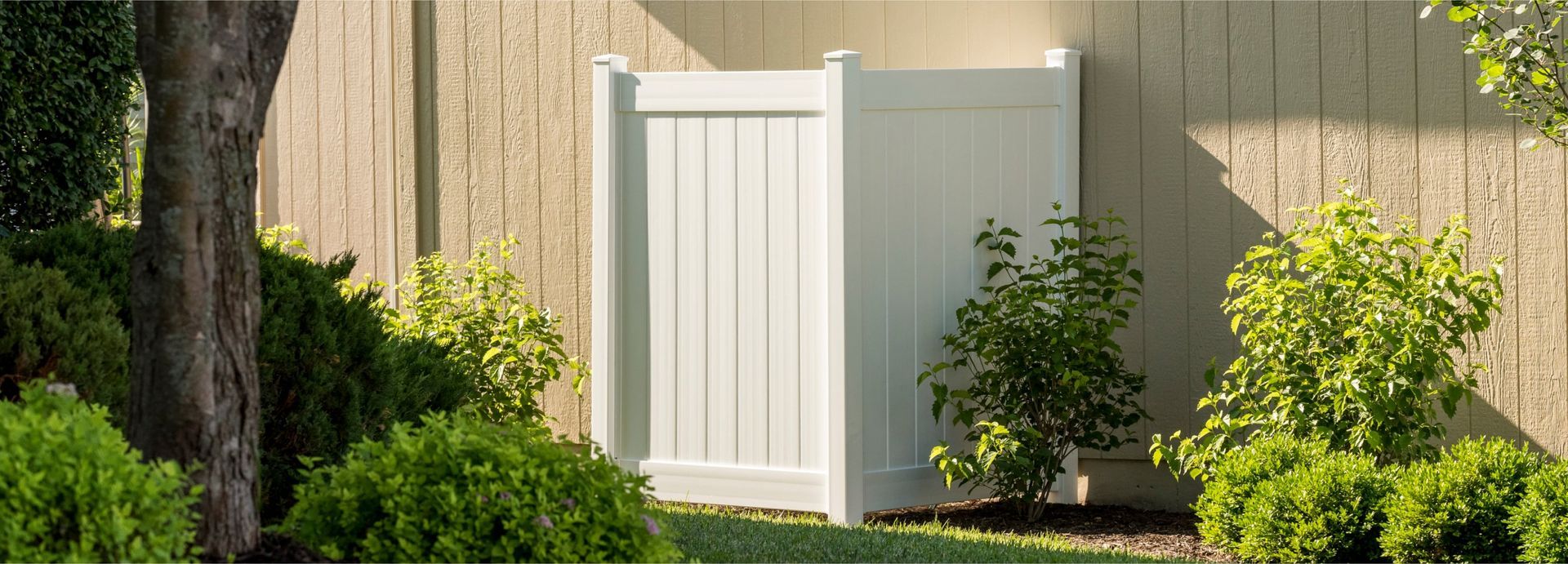 White fence with vertical panels stands in front of a neutral textured wall and is surrounded by greenery and a tree.