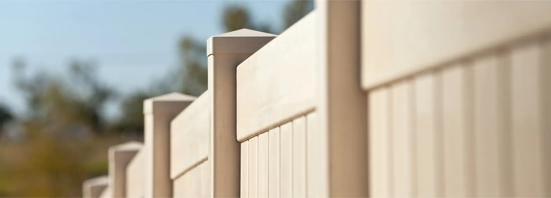 A close-up of a beige vinyl fence against a blurred background of trees and sky.