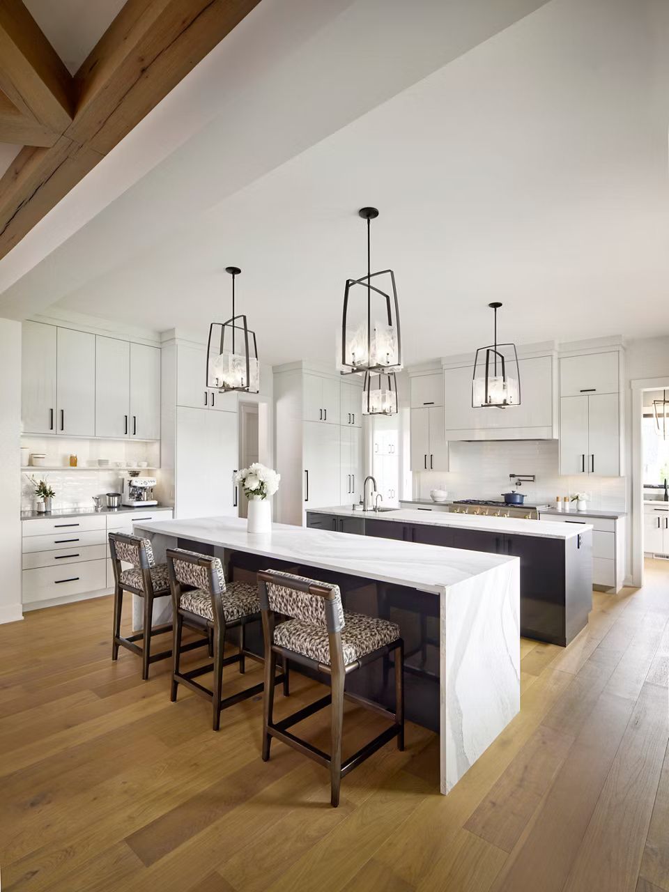 Modern kitchen with two marble-topped islands, dark cabinets, white walls, and wooden floors. Three pendant lights hang above.